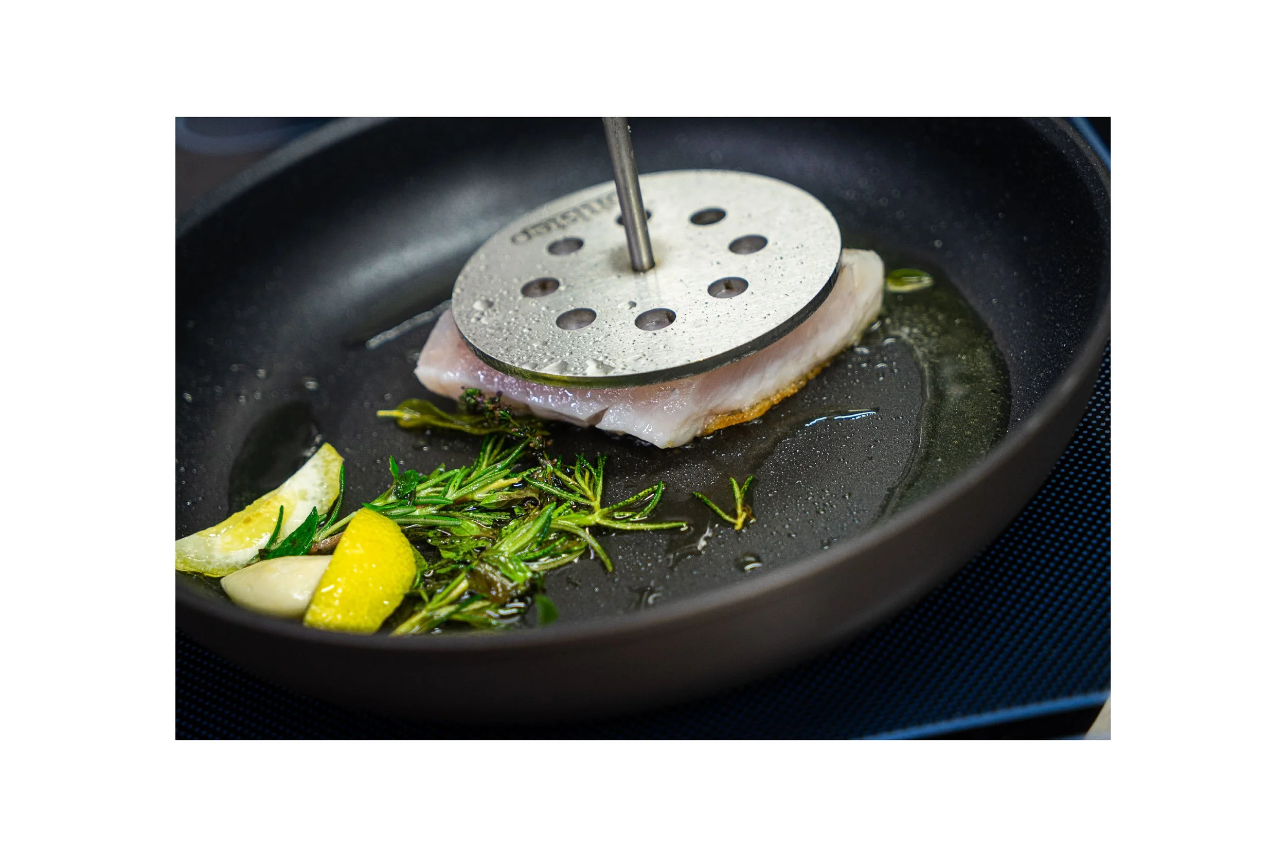 Cooking fish fillet with lemon, garlic, and herbs in a skillet with a metal fish grilling rack.