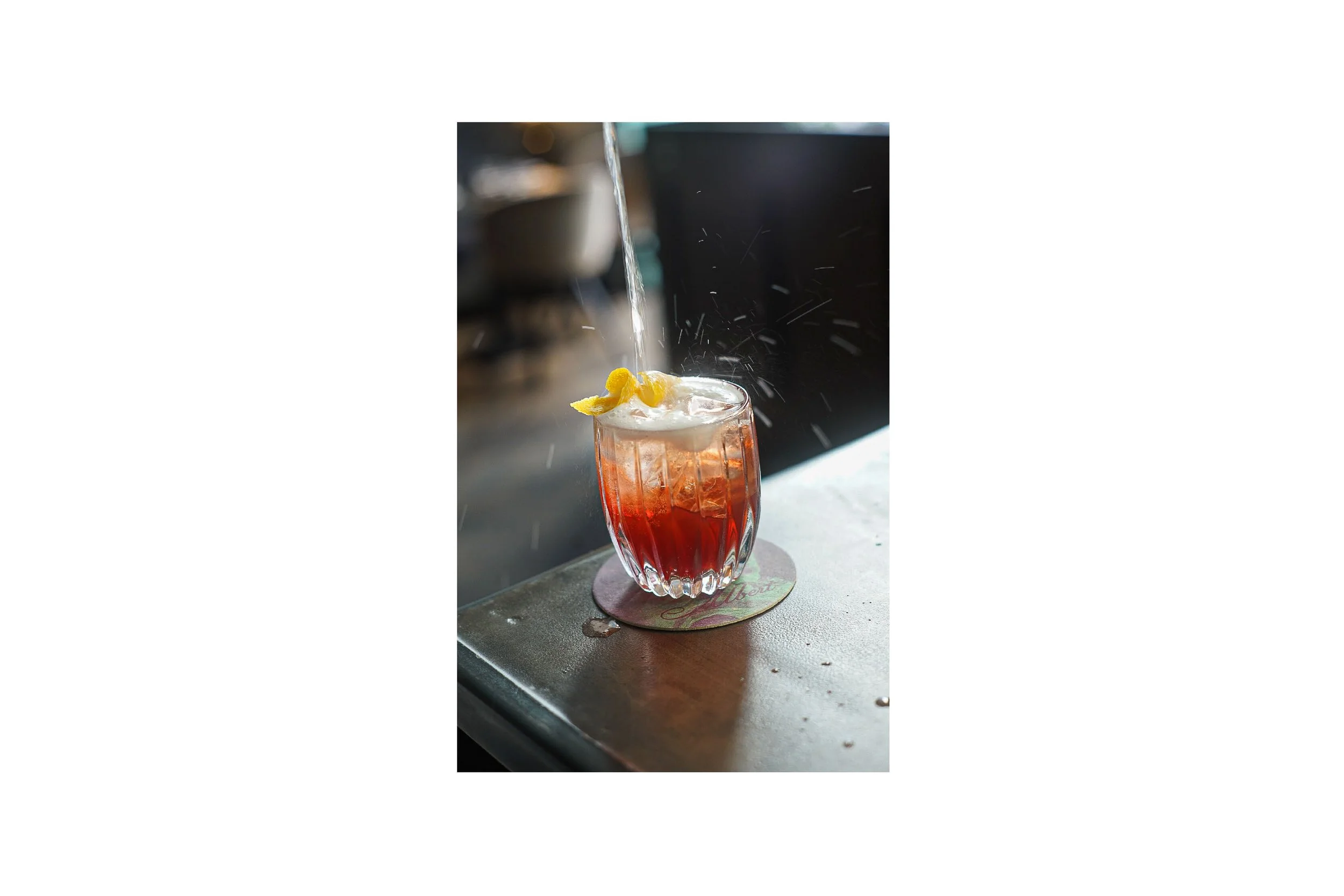 A cocktail glass with a red drink topped with foam and lemon peel, with a splash of water falling into it, placed on a coaster on a bar counter.
