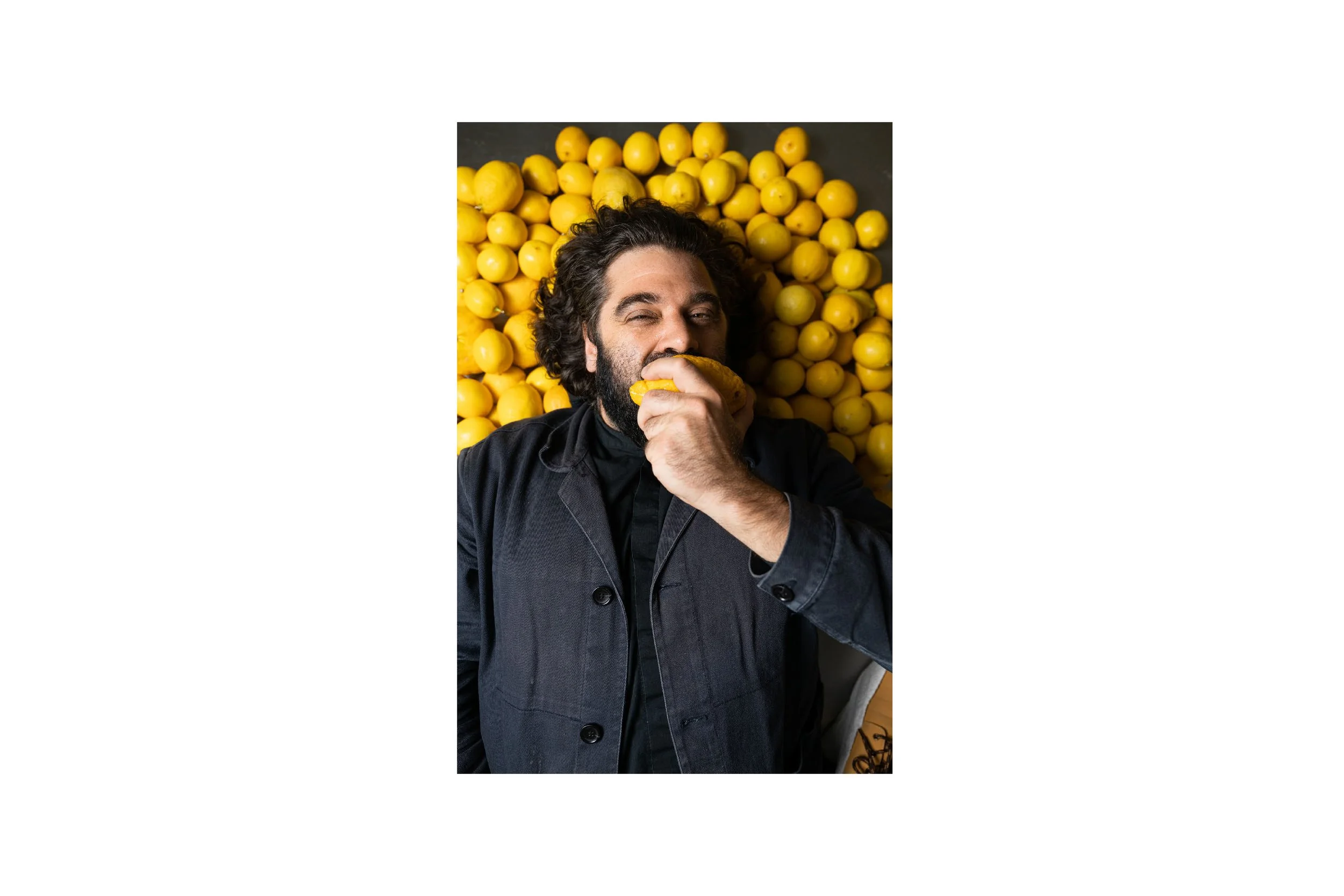 A man with dark curly hair and a beard, wearing a dark jacket, is lying on a surface covered with yellow lemons. He is holding a lemon to his mouth and appears to be about to take a bite, with one eye closed and a playful expression.