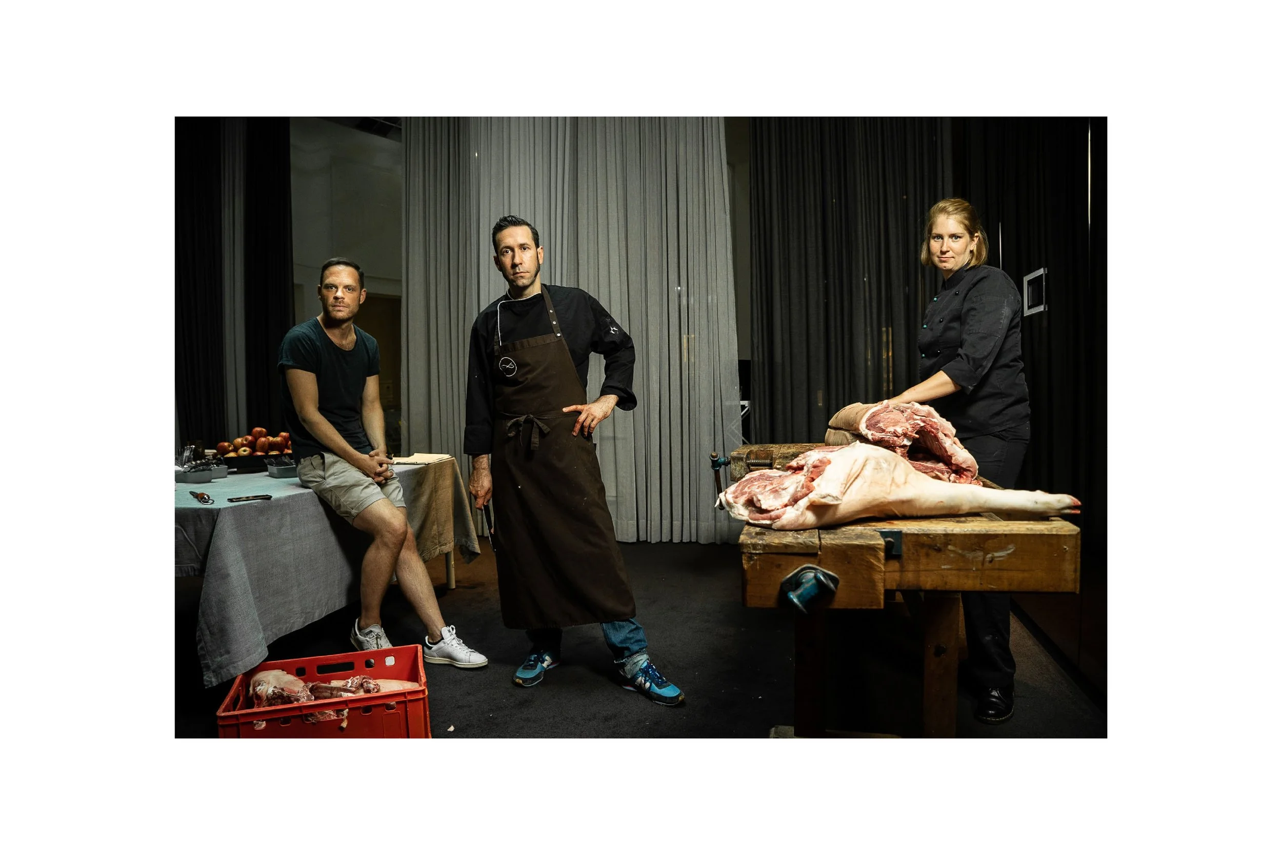 Three people in a room with butchering tools and large animal carcasses on a wooden table, with butchered meat and an apple display to the side.