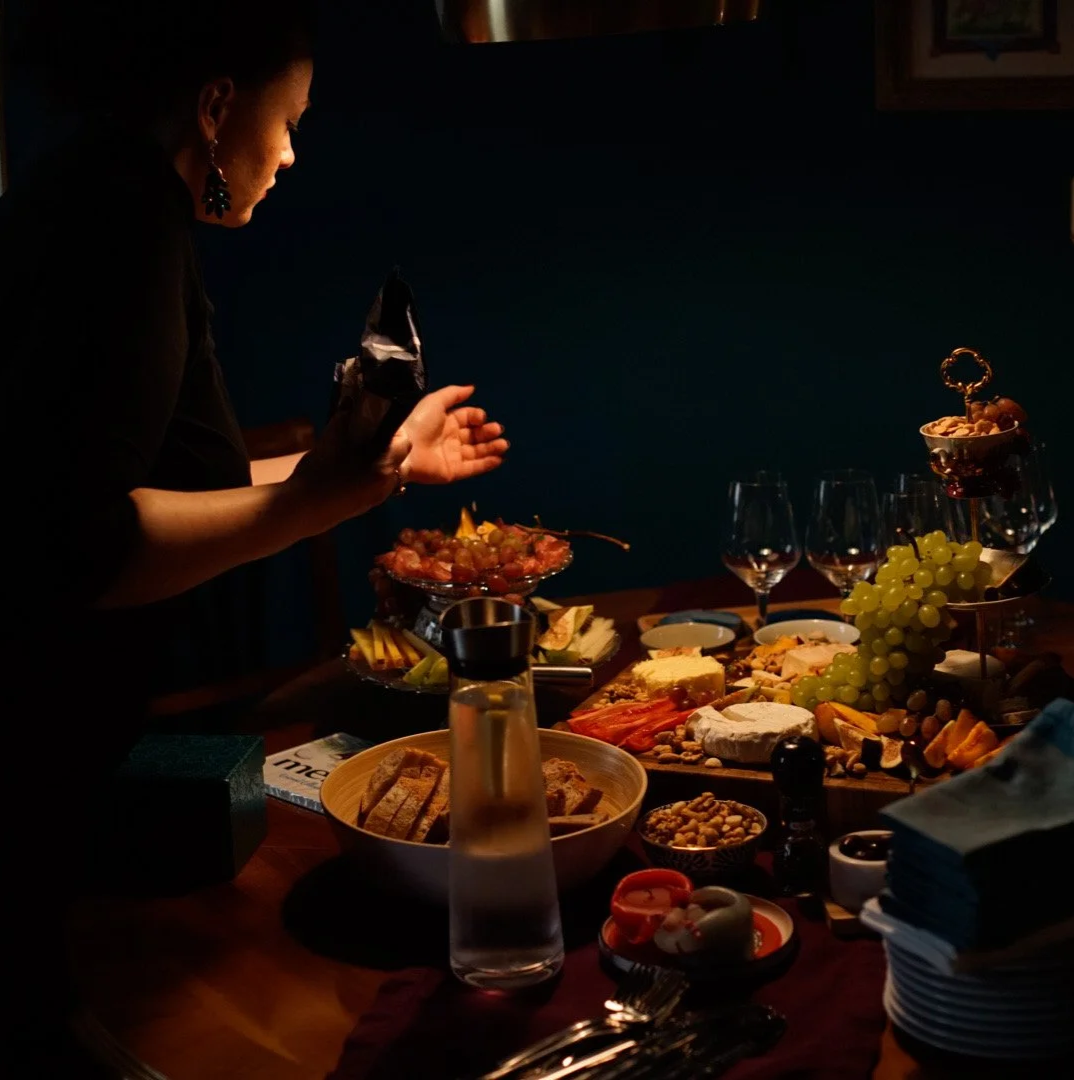 Woman arranging an abundant dinner table with cheese, fruit, glasses and tableware in warm, low light.