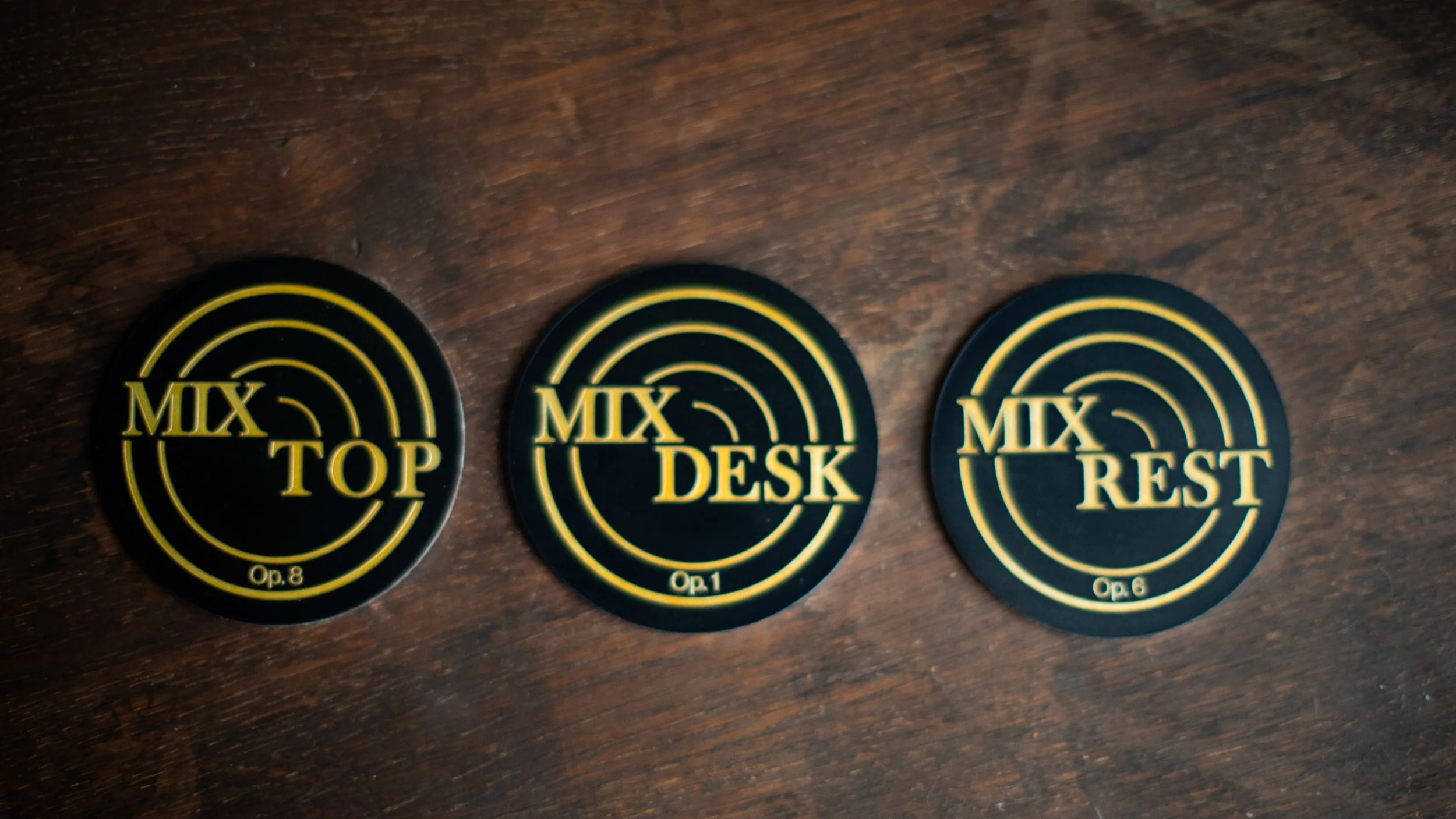 Three circular black and yellow coasters labeled 'MIX TOP', 'MIX DESK', and 'MIX REST' arranged on a dark wooden surface.