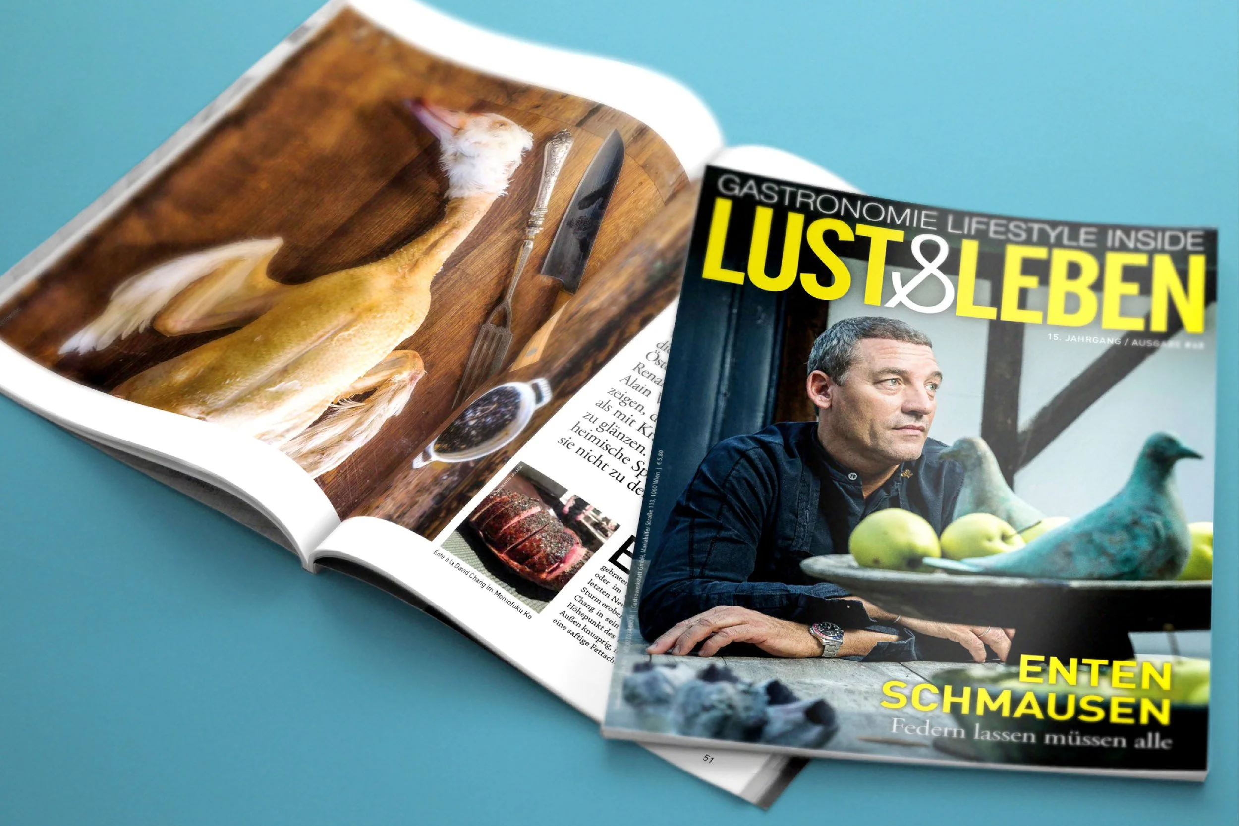 A magazine titled "LUST & LEBEN" with a man sitting at a table featuring a bowl of apples and ceramic birds on the cover, and an open magazine with a photograph of a dead fish and kitchen utensils on the purple background.