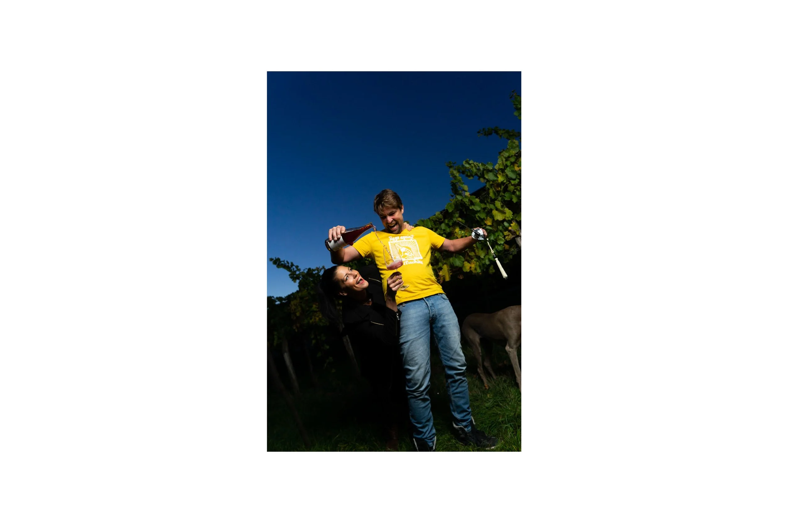 Two friends celebrating outdoors at night, pouring beer into glasses, with a dog nearby and a clear dark sky in the background.
