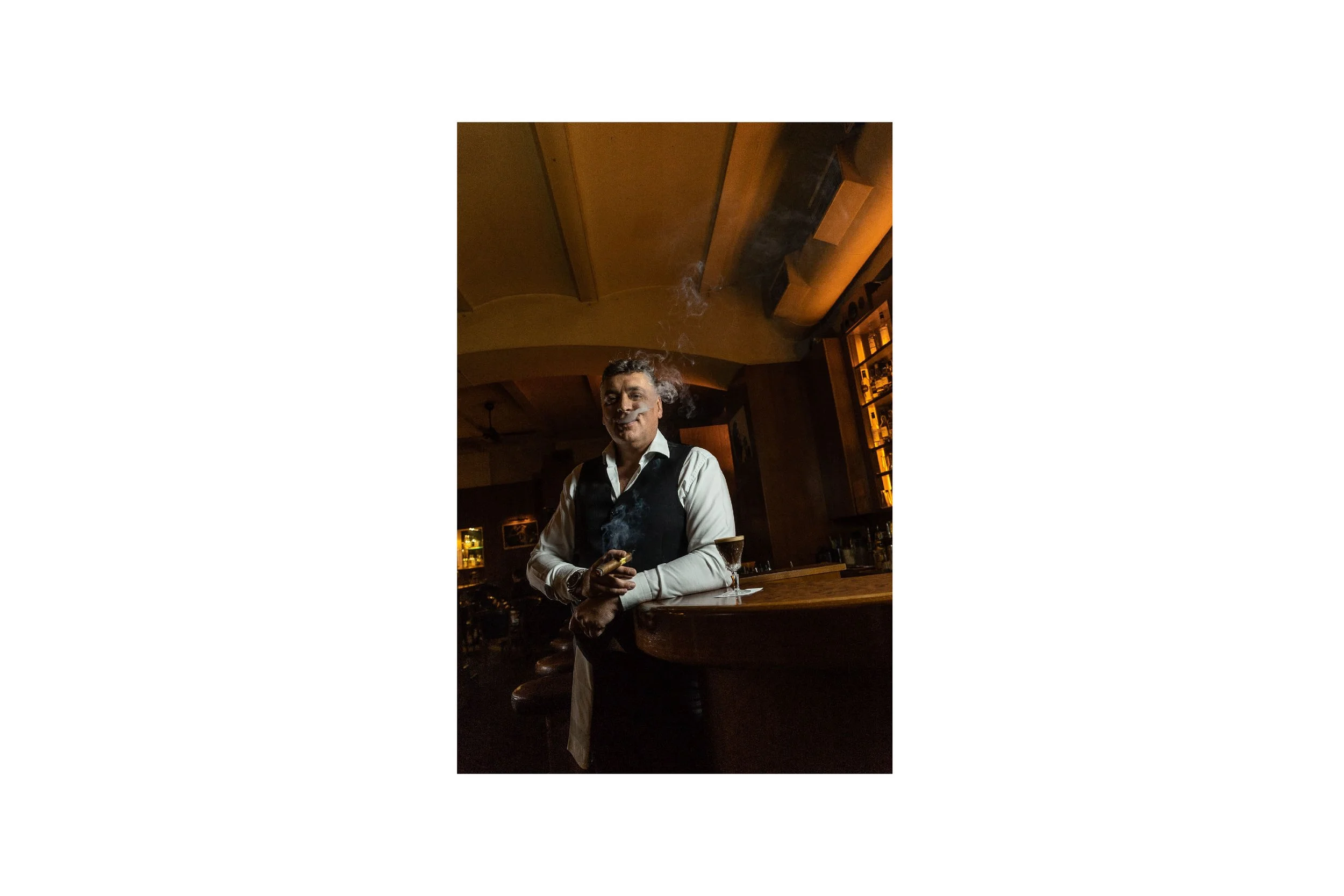 A bartender in a bar, wearing a white shirt and black vest, standing behind a wooden counter with a cocktail glass nearby, amid a warm and dimly lit atmosphere.