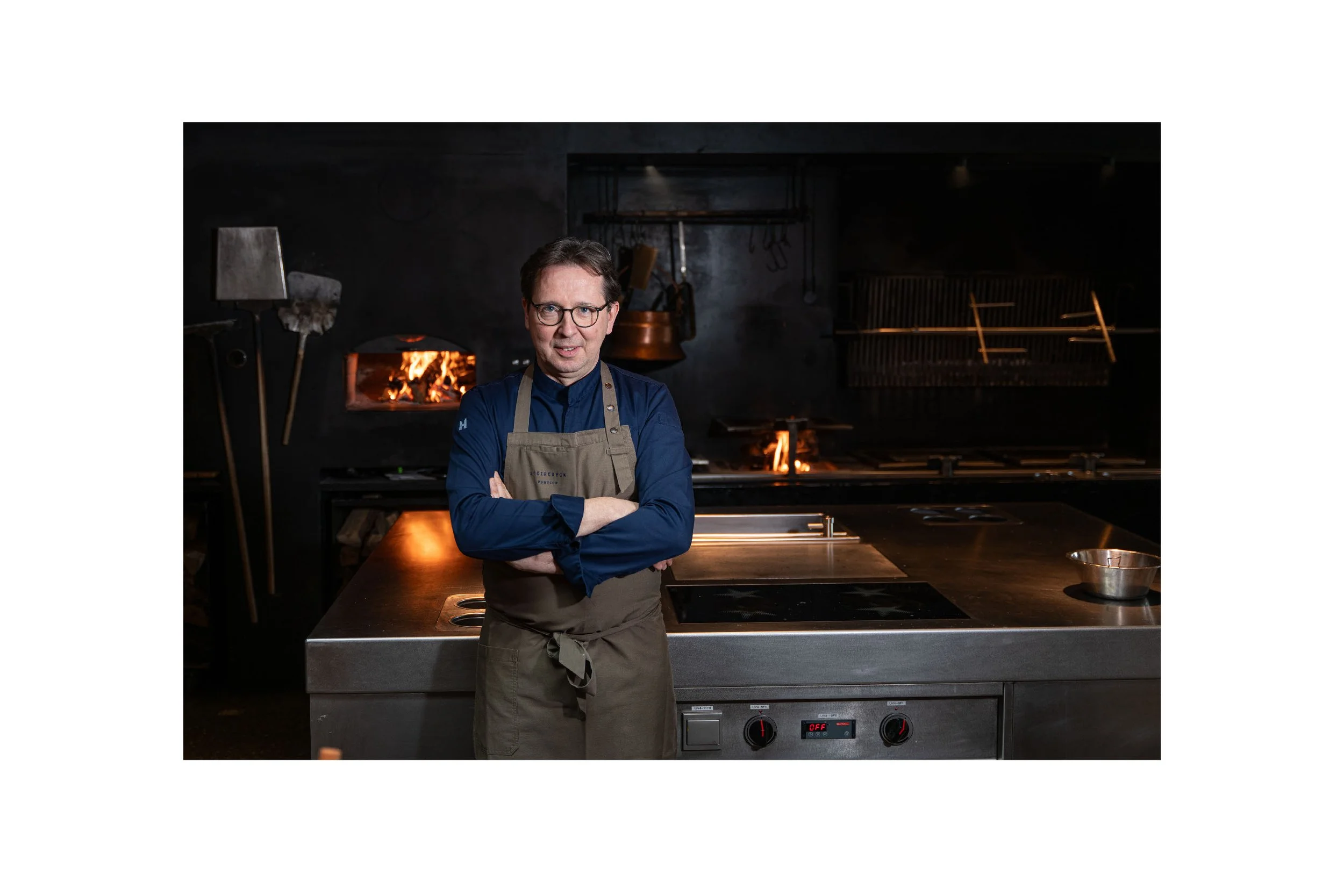 A man in a blue shirt and apron standing with arms crossed in a professional kitchen with a fire burning in the oven behind him.