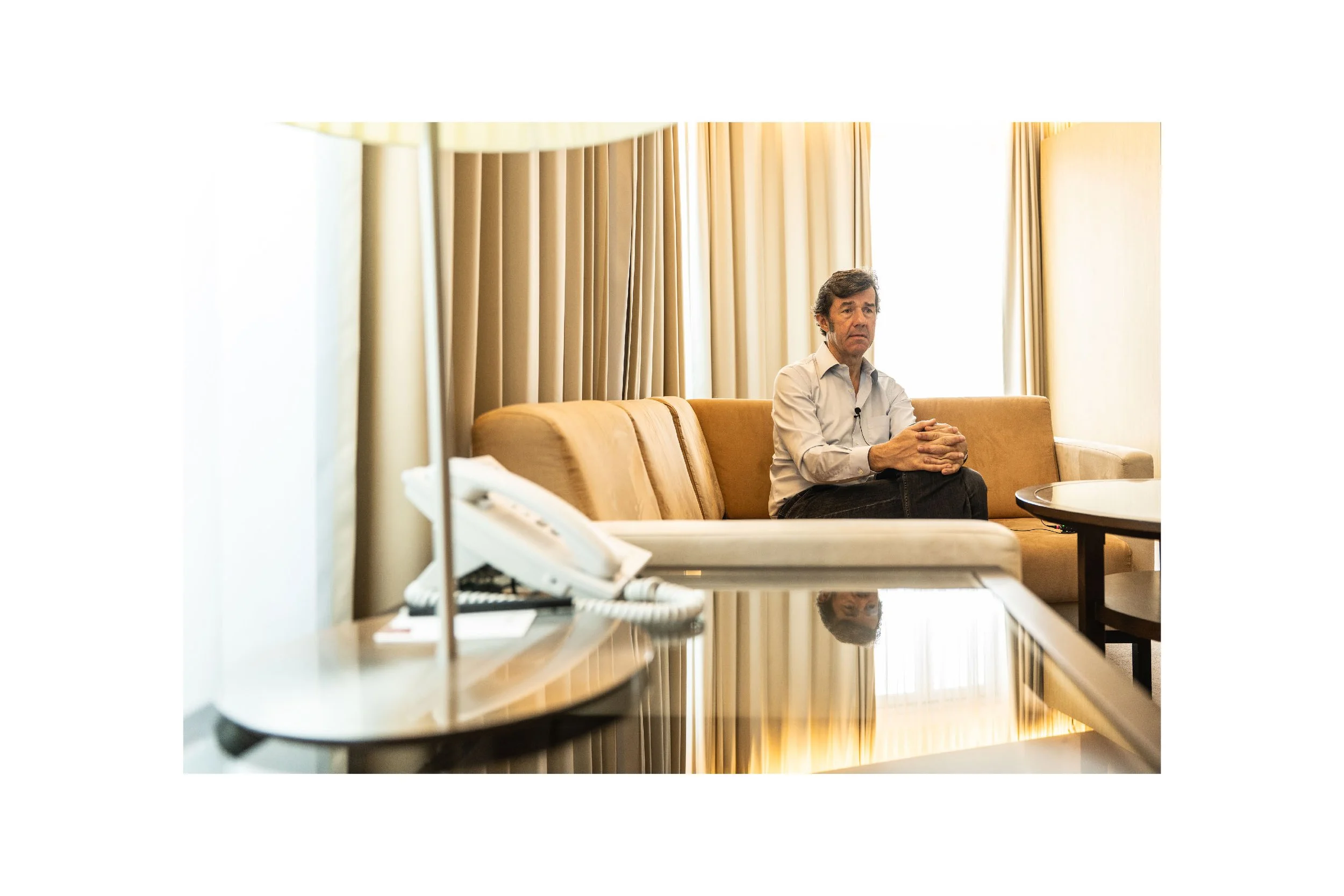A man sitting on a beige sofa in a hotel room with curtains behind him, looking pensively forward.