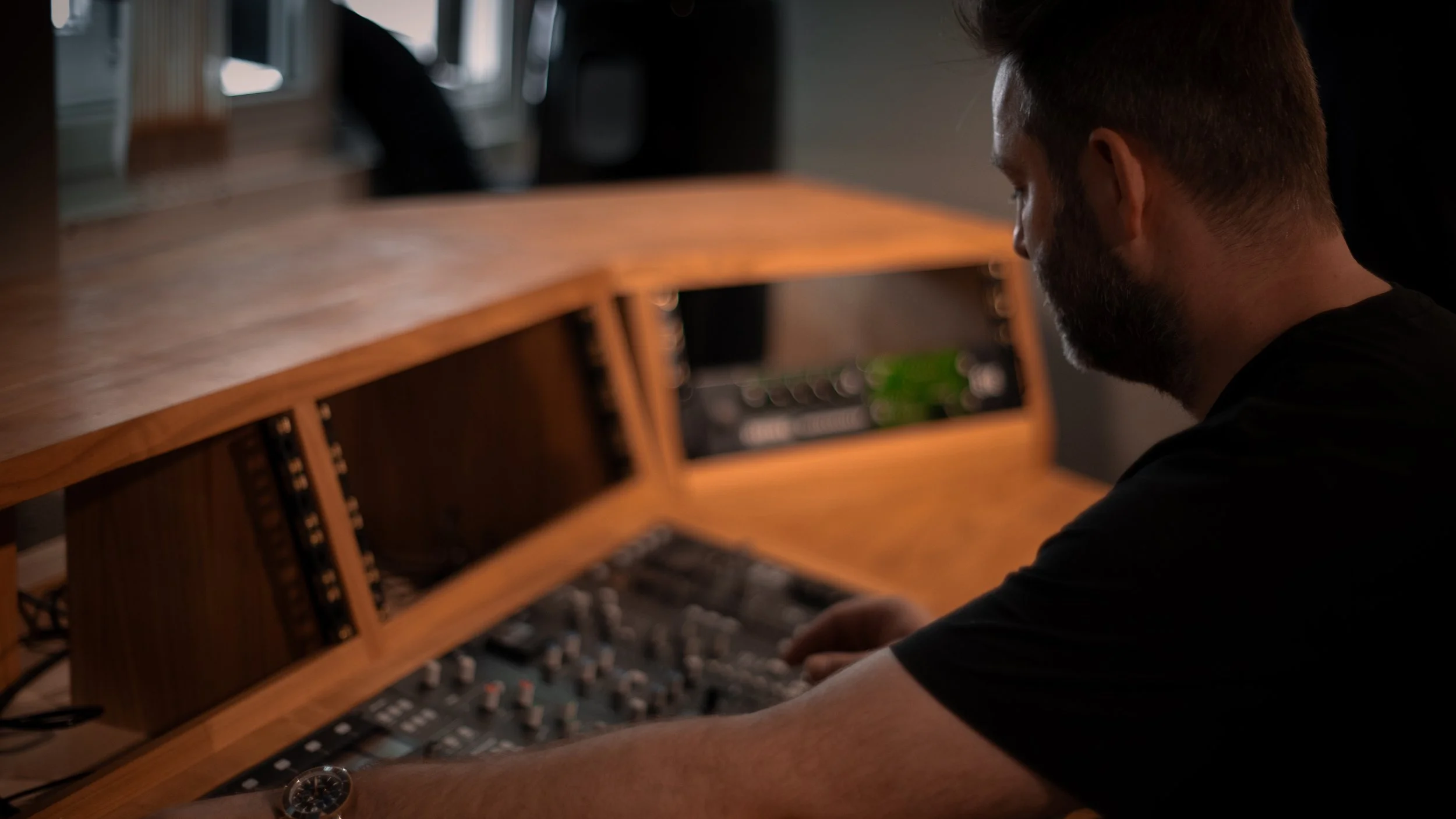 A man with a beard and short hair is sitting at a wooden music studio desk, working with audio equipment.
