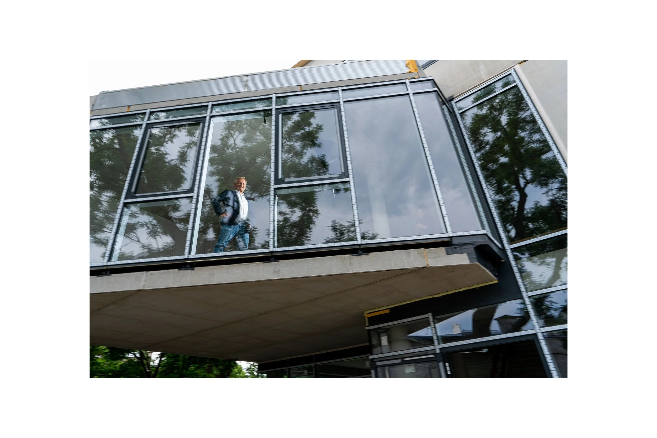 A man standing inside a building with glass walls, looking out through the reflection of trees.