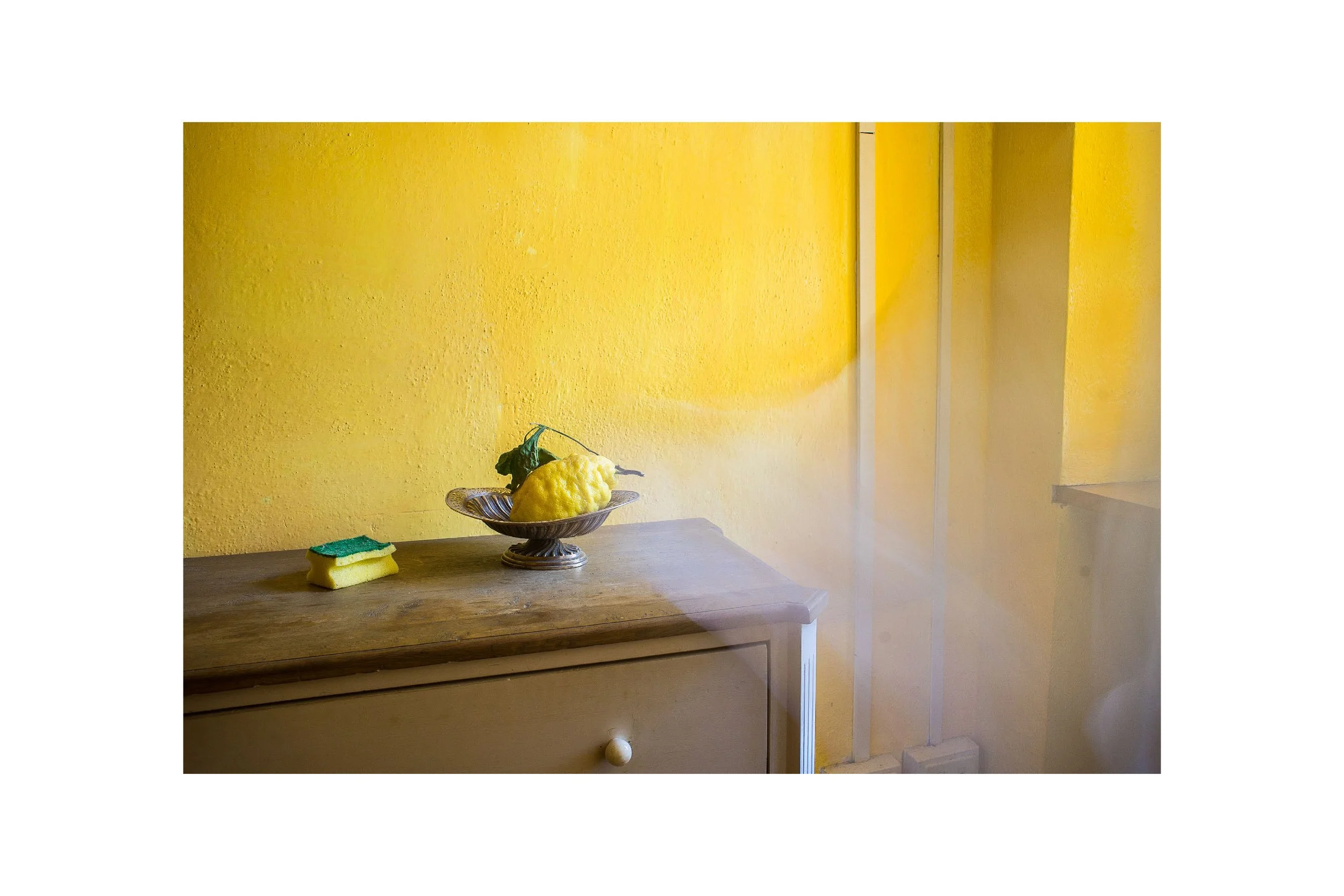 A wooden dresser with a yellow decorative fruit, a sponge, and a bowl on top, against a yellow wall.