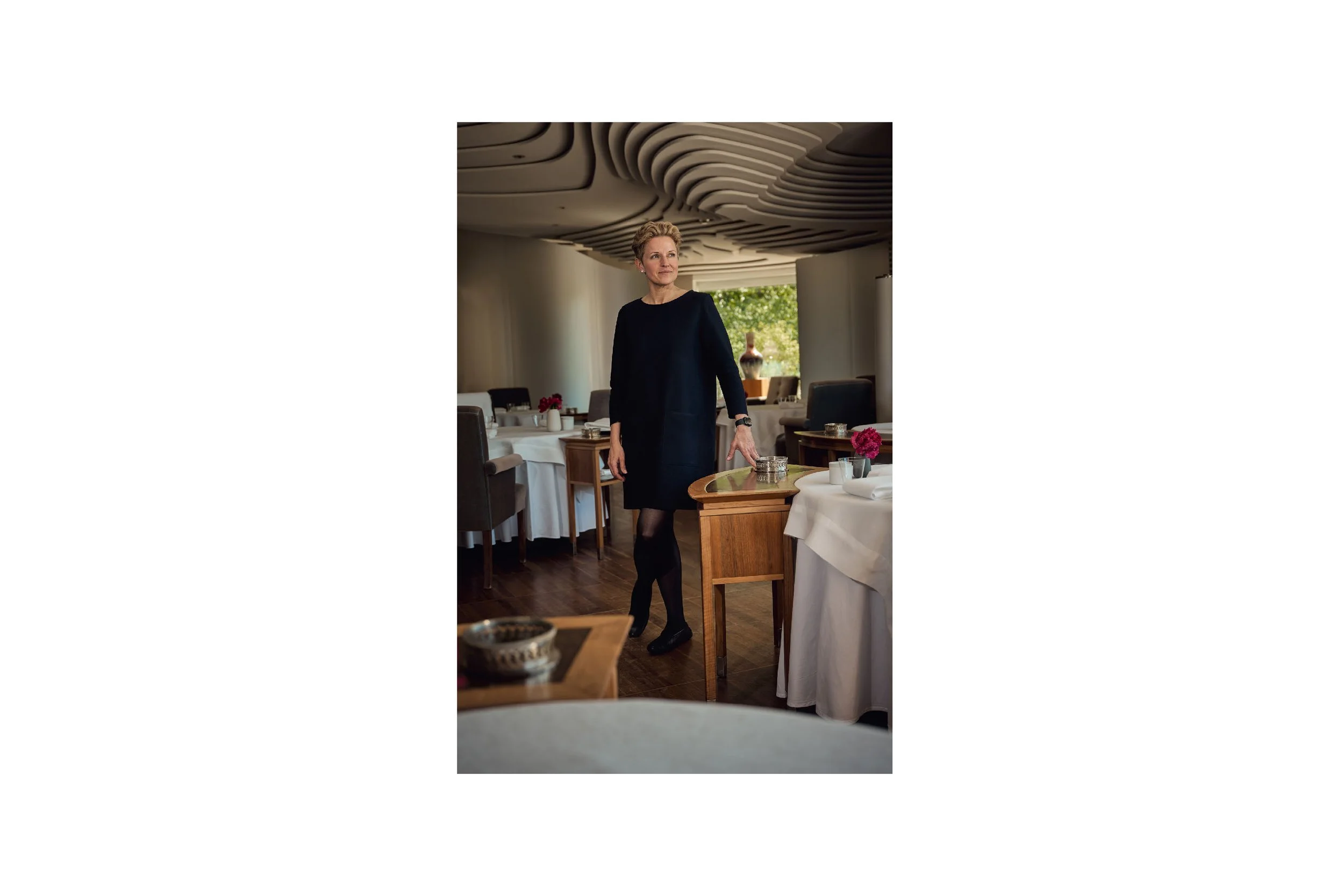 A woman in a black dress and tights standing in a restaurant dining area with white tablecloths, pink flowers, and wooden furniture, looking off to the side.
