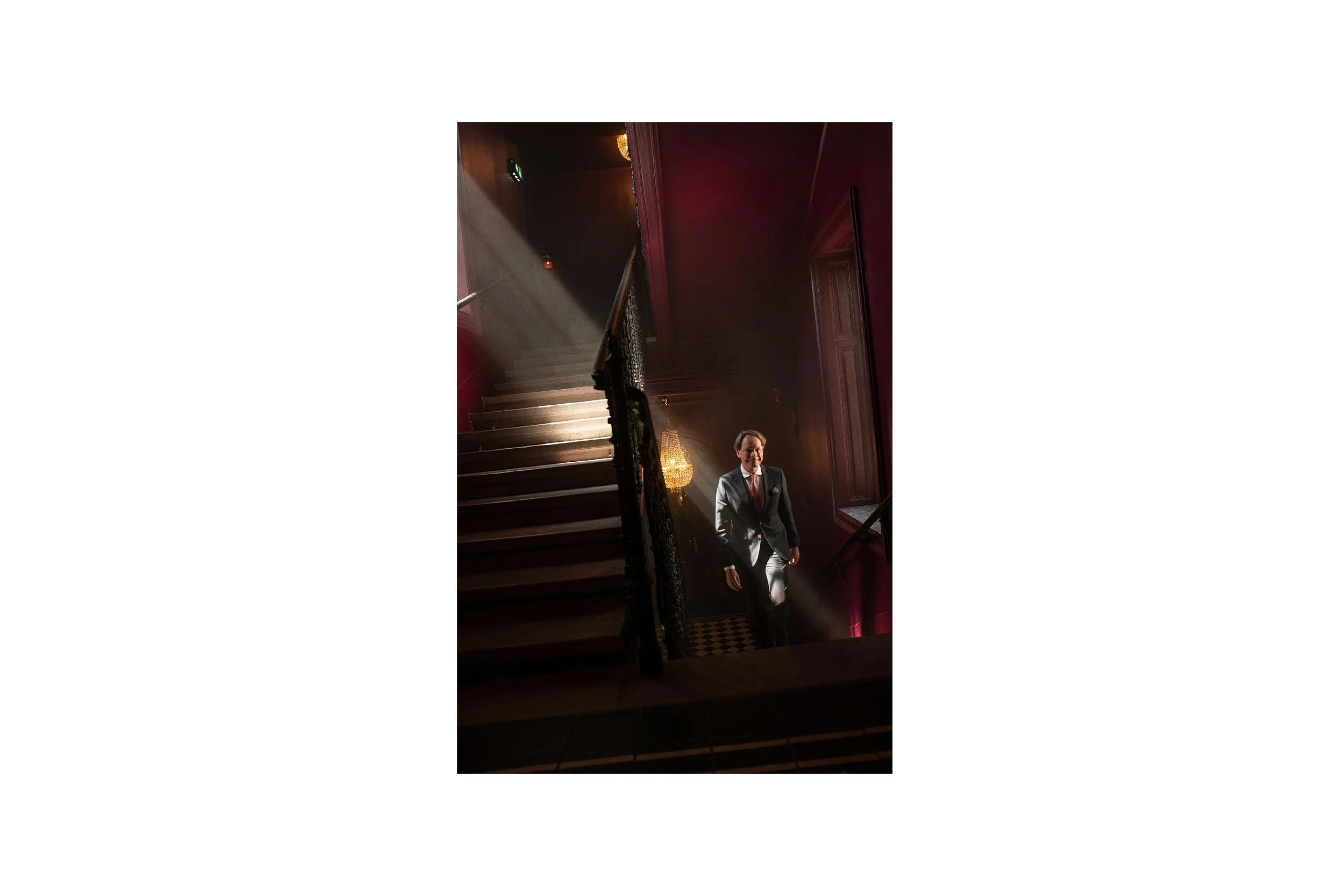 A man in a gray suit with a red tie walking down a staircase in a dimly lit, elegant interior with dark red walls and a chandelier.