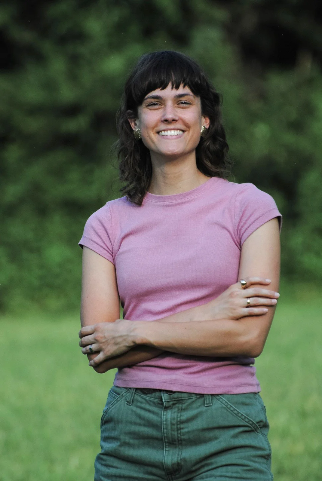 Journalist and writer Sarah Melotte stands in a pink tee shirt and green pants with her arms crossed against a blurry green background outside.