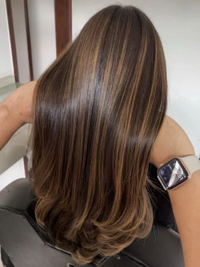 A woman with long, shiny, light brown hair sitting on a black chair, wearing a smartwatch on her left wrist.