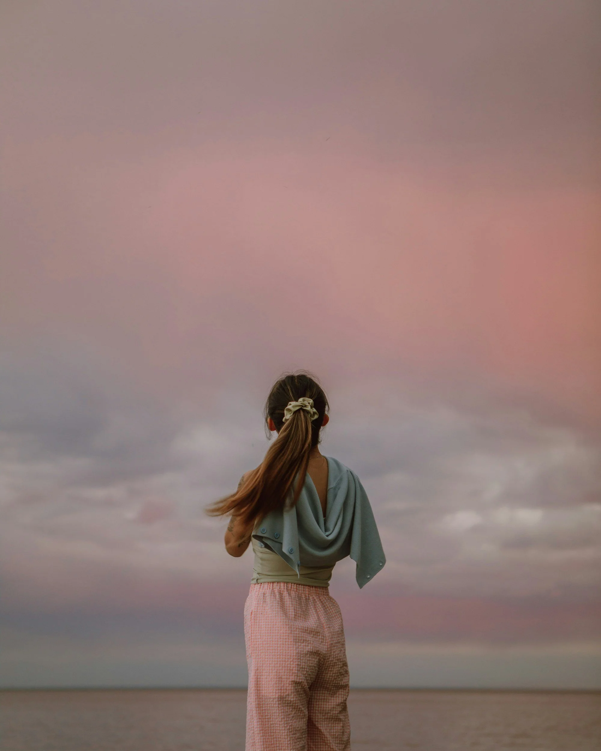 A girl with long hair tied back, standing by the water, facing a colorful sunset sky.