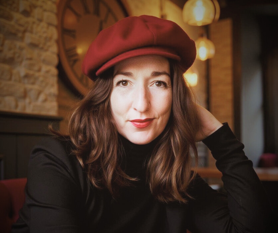 A woman with brown hair wearing a red beret and black top, sitting in a warmly lit cafe with brick walls and large wall clock.
