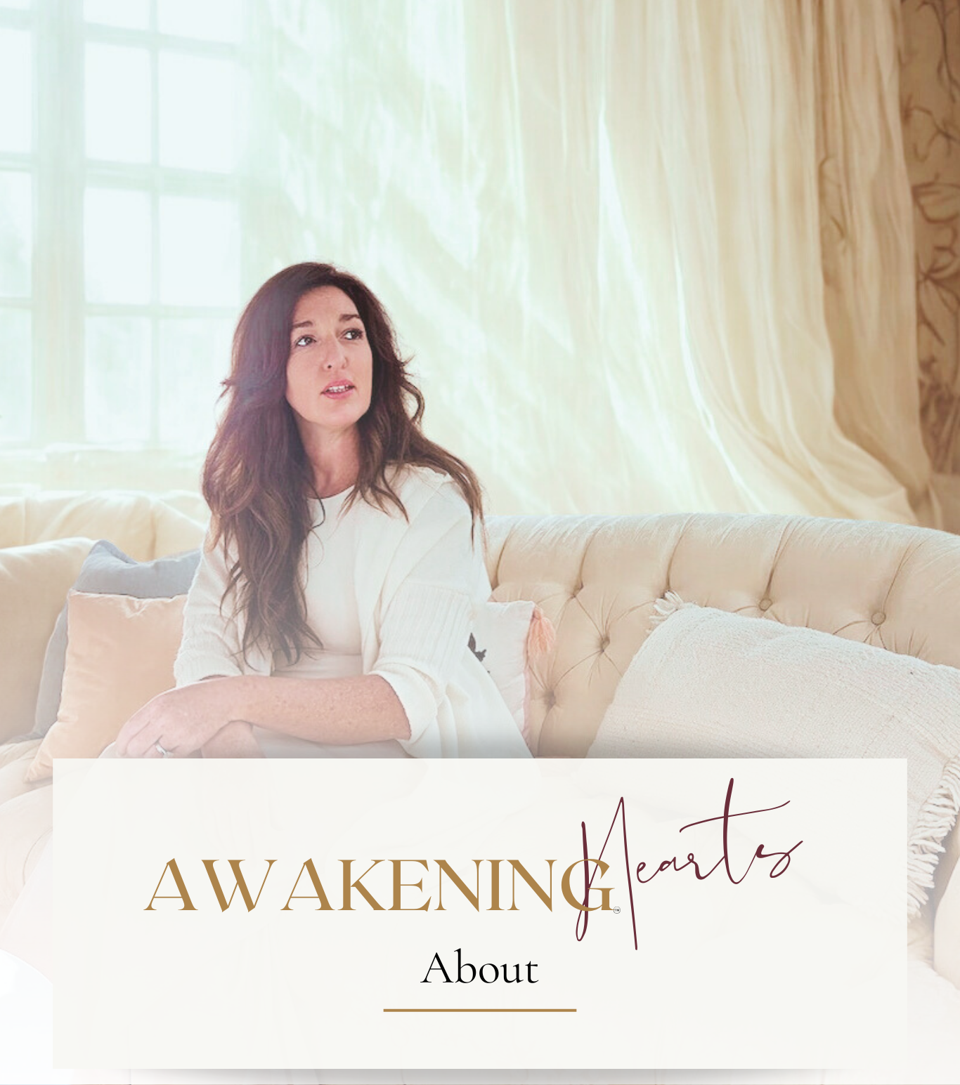 A woman with long wavy brown hair sits on a cream-colored tufted sofa in a room with large windows and beige curtains, looking thoughtful.