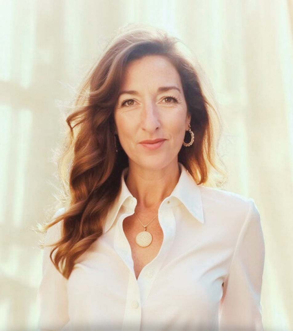 A woman with long, wavy brown hair wearing a white button-up shirt, gold earrings, and a gold pendant necklace, standing in front of a soft, light-colored background.