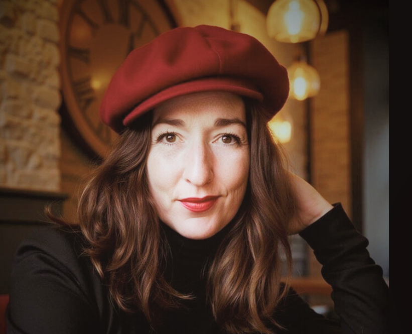 Woman with wavy brown hair wearing a red beret and black turtleneck in a cozy cafe or restaurant with warm lighting and a large wall clock in the background.