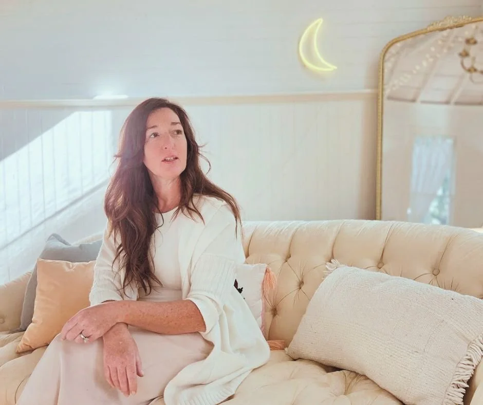 A woman with long wavy brown hair sitting on a cream-colored tufted sofa in a light, cozy room with a moon-shaped wall decoration and a large mirror.