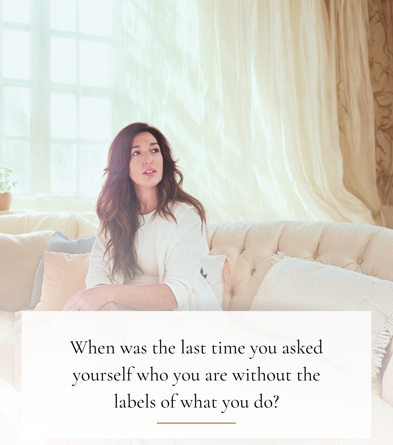 A woman with long brown hair sitting on a cream-colored sofa, looking thoughtful, with a window and sheer curtains in the background.