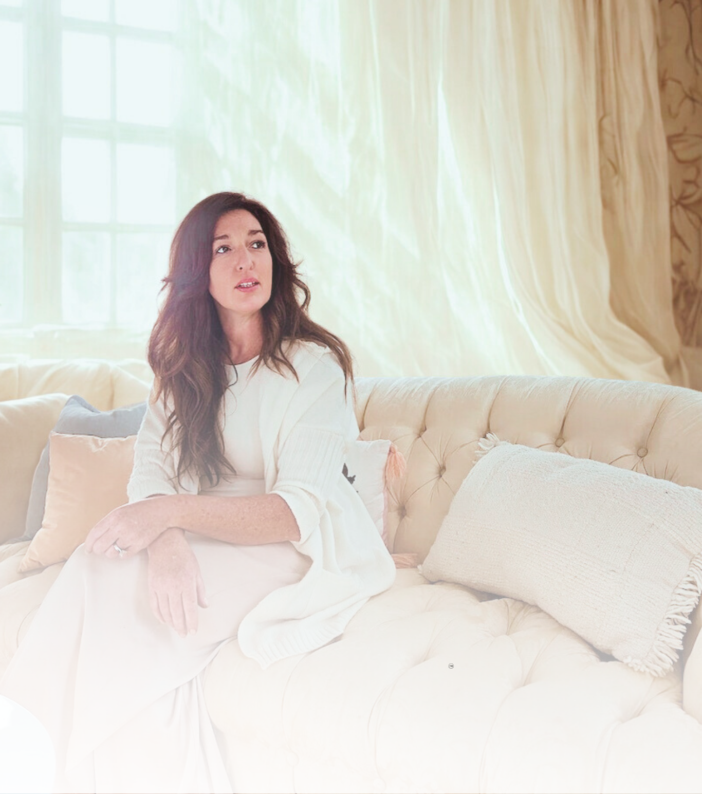 A woman with long dark hair sitting on a beige tufted sofa in front of large window with cream curtains. She is wearing a white dress or top, looking slightly to her right with a thoughtful expression.