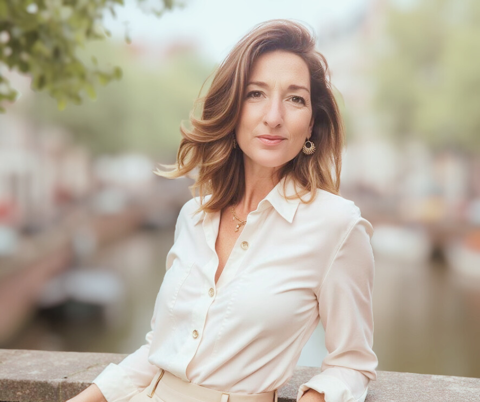 A woman with shoulder-length brown hair stands outdoors near a body of water, wearing a cream-colored blouse and looking at the camera.