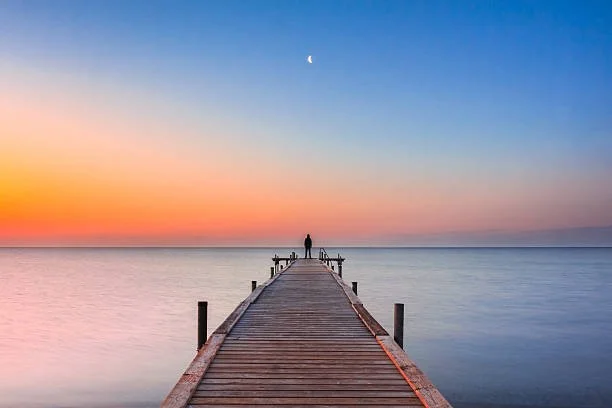 A person standing on a wooden pier extending into a calm body of water during sunset with colorful sky and a crescent moon.