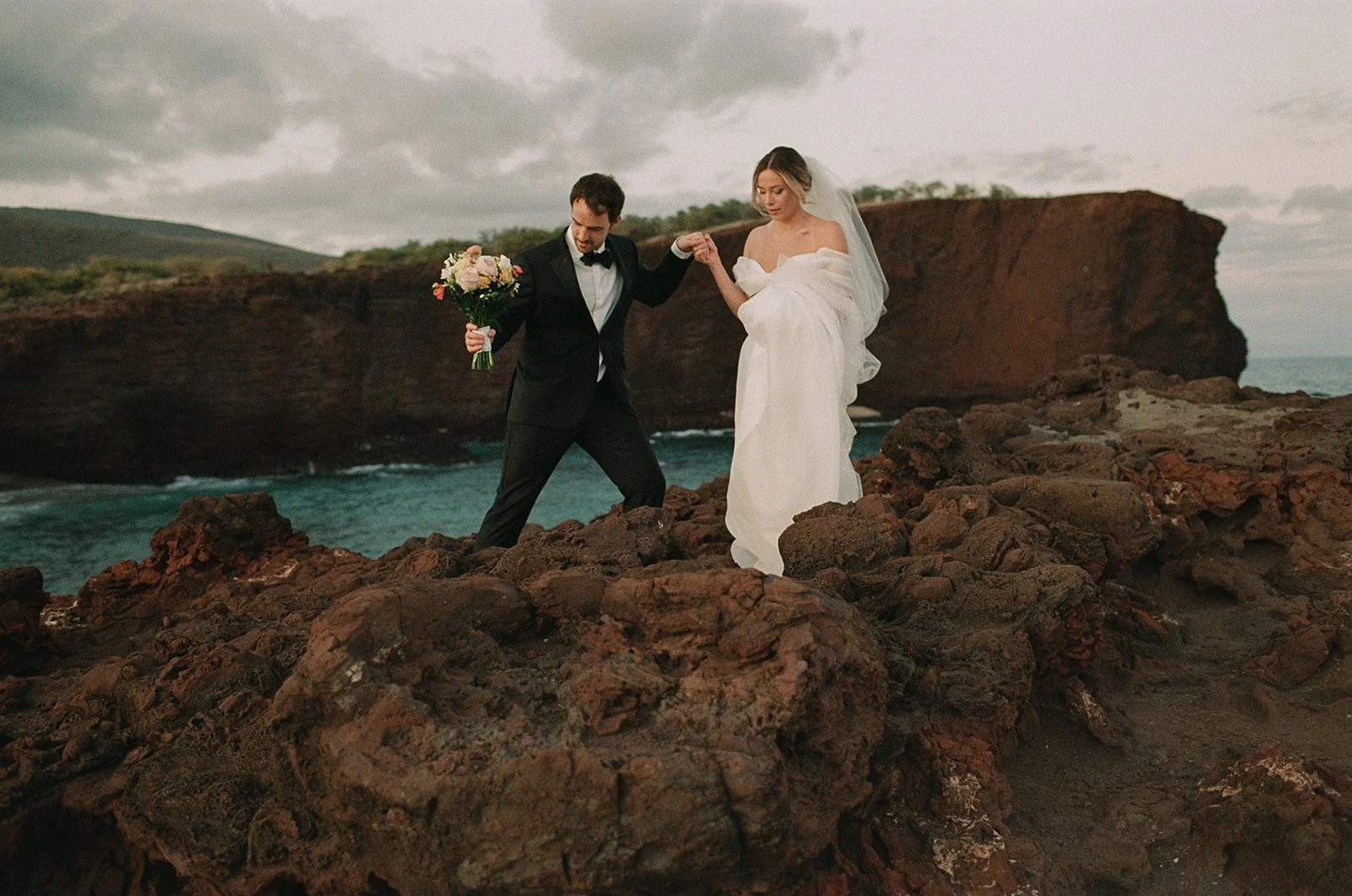 A bride and groom on a rocky cliff, with the groom holding a bouquet of flowers and helping the bride walk, both dressed in wedding attire, against a backdrop of cliffs and cloudy sky.