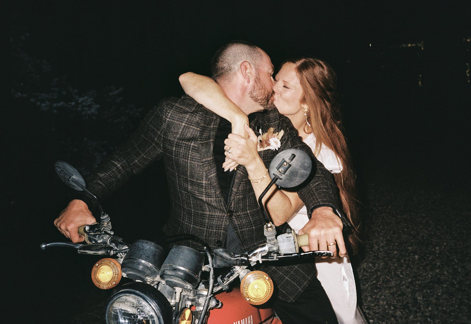 A couple sharing a kiss on a motorcycle at night, dressed in stylish clothing, with dark background and minimal lighting.
