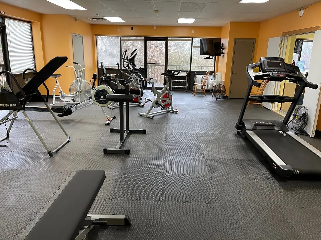 Empty indoor gym with exercise equipment including a treadmill, stationary bikes, and elliptical machines, with windows and a balcony in the background.