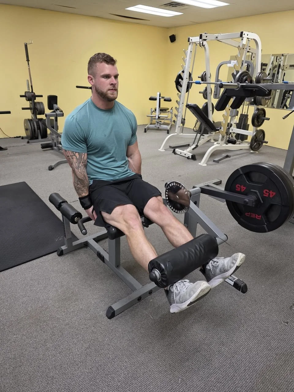 A man in a gym sitting on a hyperextension bench with weights and gym equipment in the background.