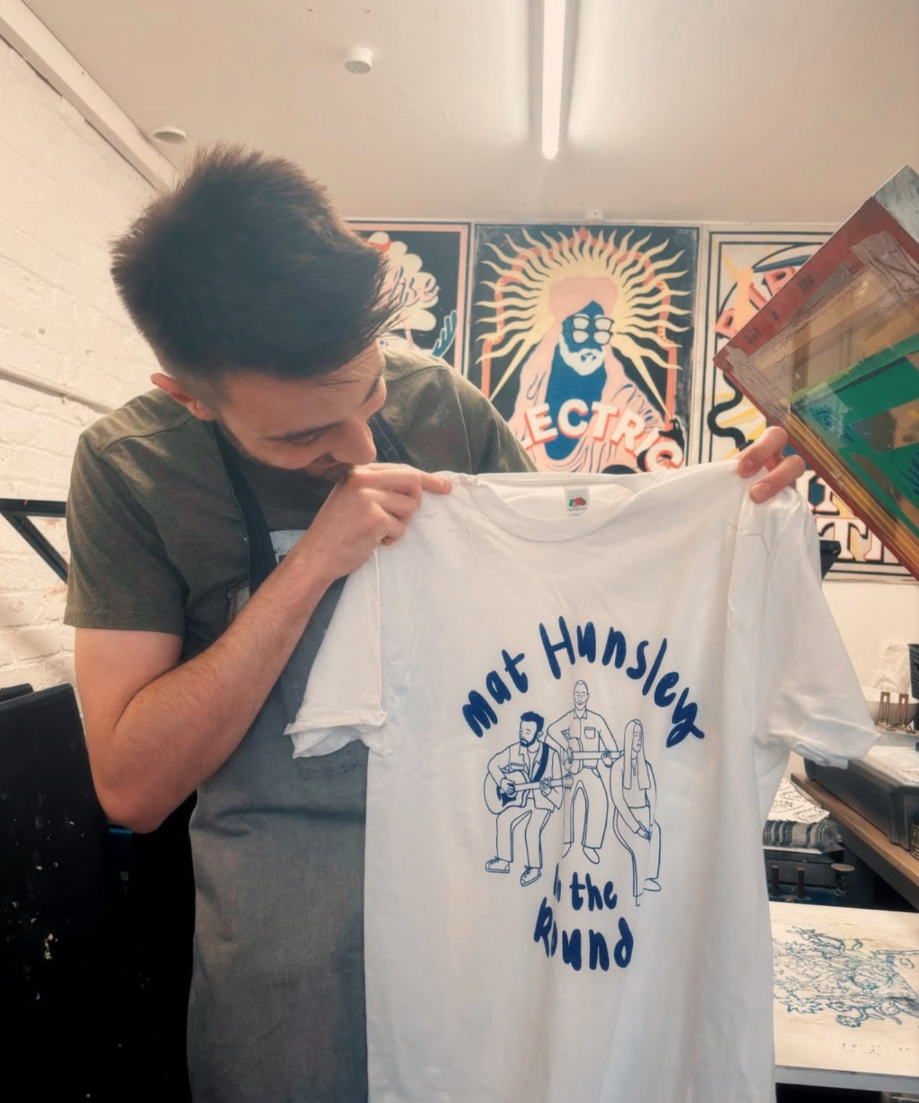 A person with dark hair looking at a white T-shirt with blue illustration and text, in a room with colorful artwork on the wall behind him.