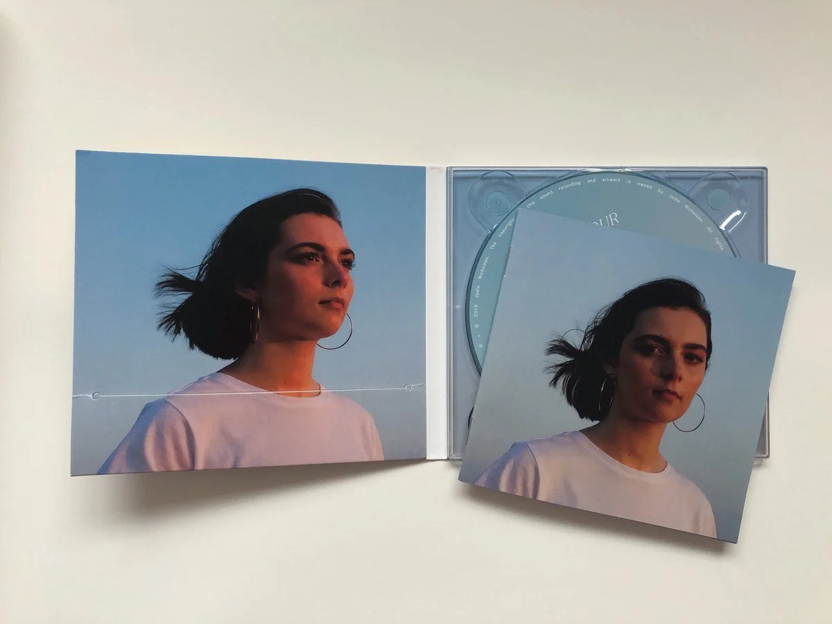 Open CD case with two photos of a woman with dark hair, wearing hoop earrings and a white shirt, against a blue sky background, one photo inside the case and one outside.