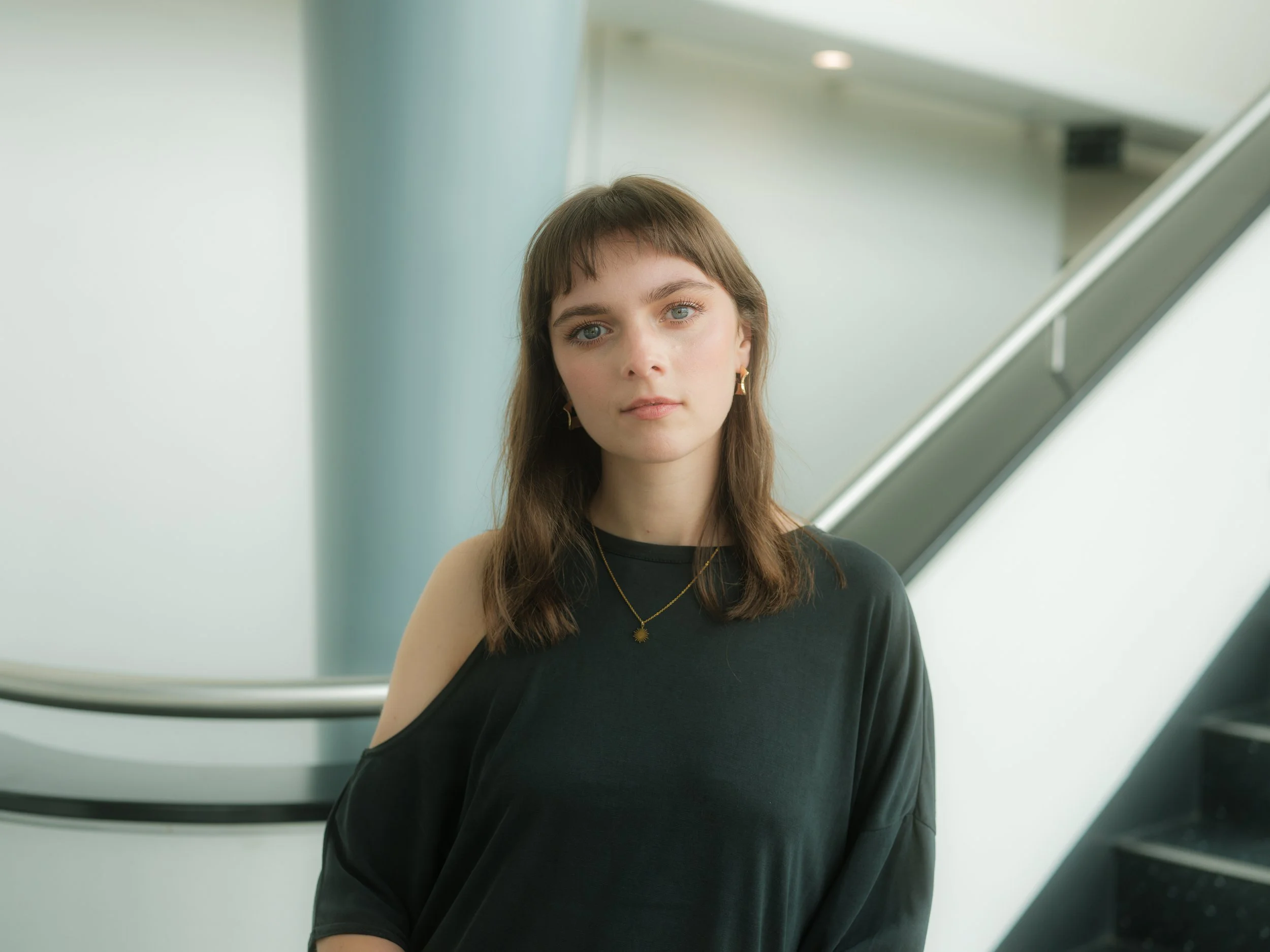 A young woman with shoulder-length brown hair, blue eyes, and fair skin standing next to a curving staircase in a modern building with light-colored walls. She wears a black off-the-shoulder top and gold jewelry, looking directly at the camera.