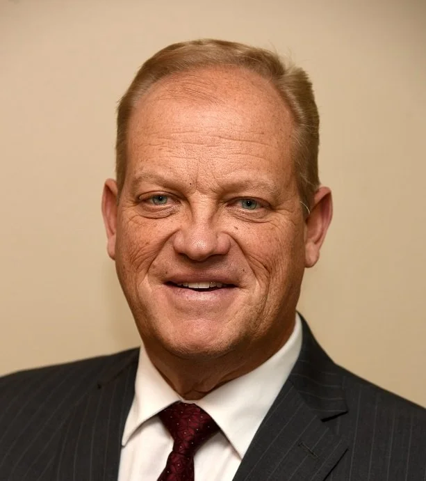 A middle-aged man with short blonde hair, blue eyes, wearing a dark pinstripe suit, white shirt, and maroon tie, smiling at the camera against a beige background.
