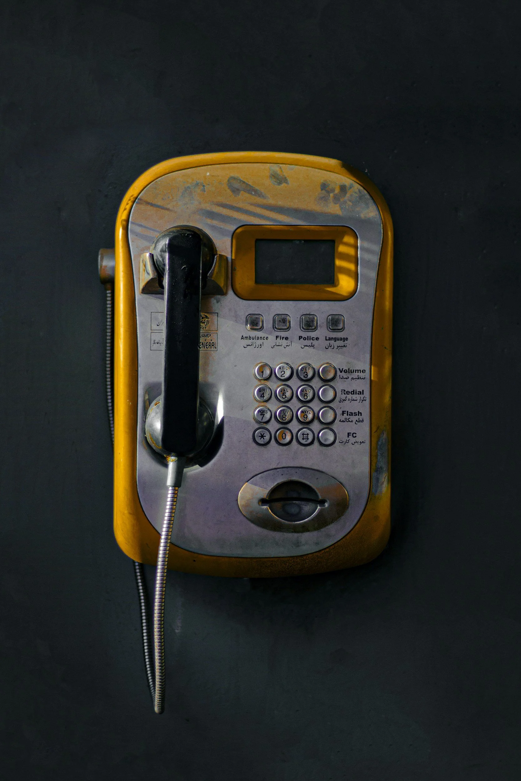 A yellow payphone mounted on a dark wall, with a black handset attached by a metal cord, and a keypad with buttons in English and Arabic.