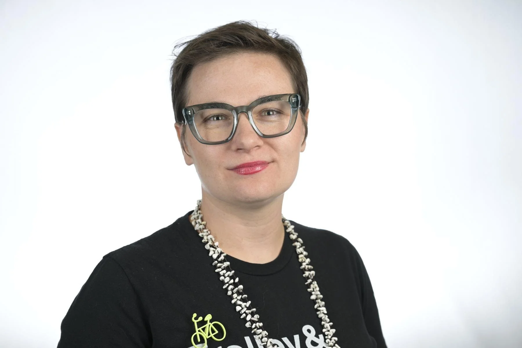 Picture of Jen Hawse. Wearing big green glasses. Smiling slightly. Big white necklace. Bike button on shirt. Short hair.