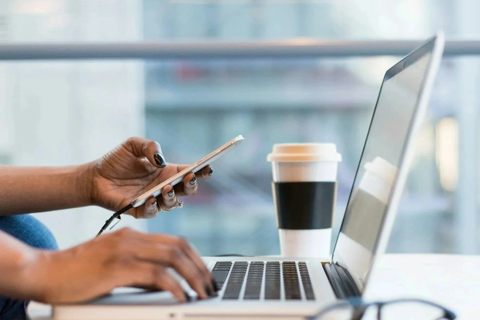 Person using a smartphone and working on a laptop with a coffee cup nearby