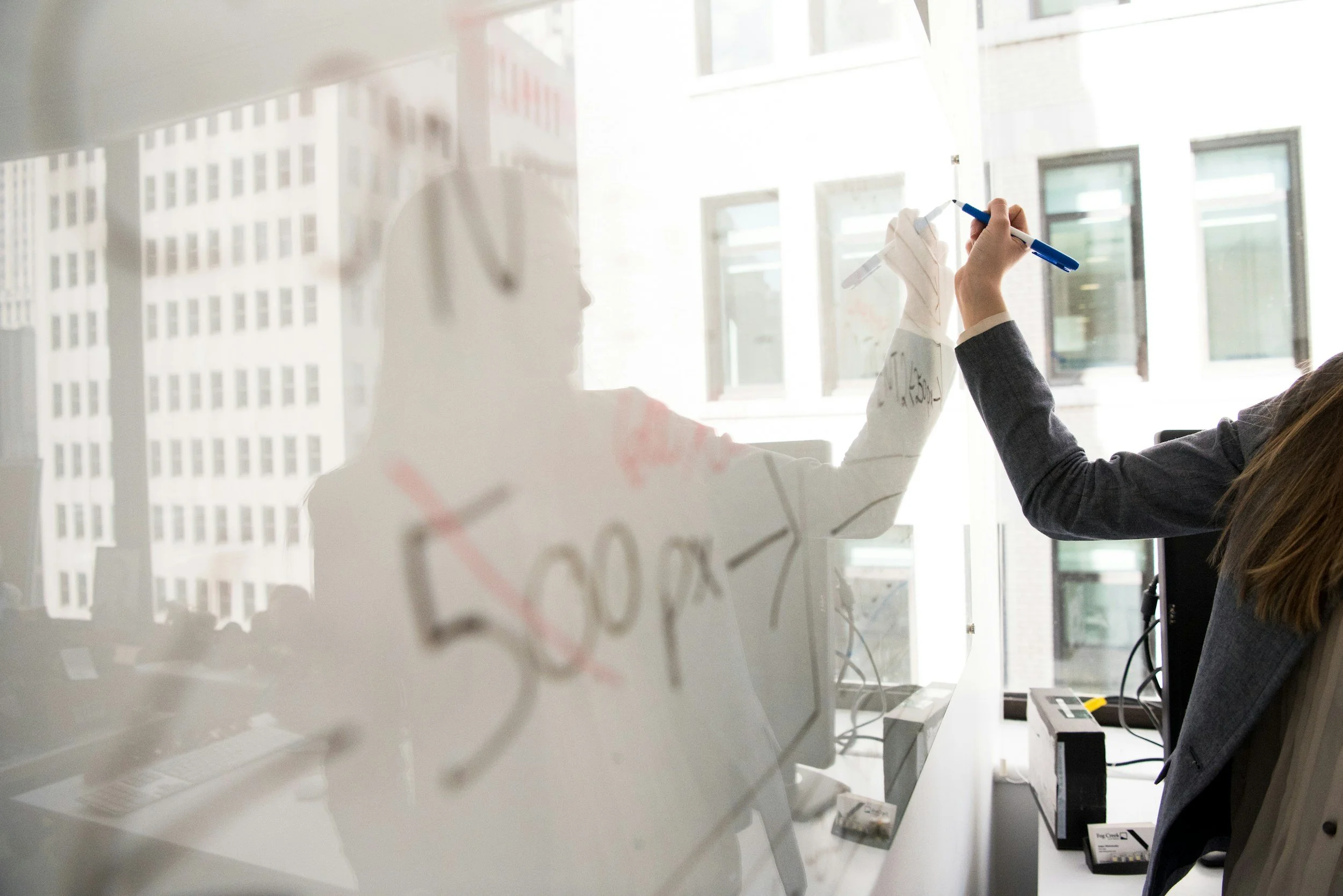 Two women are writing on a transparent glass wall with marker pens in an office, with city buildings outside the windows.