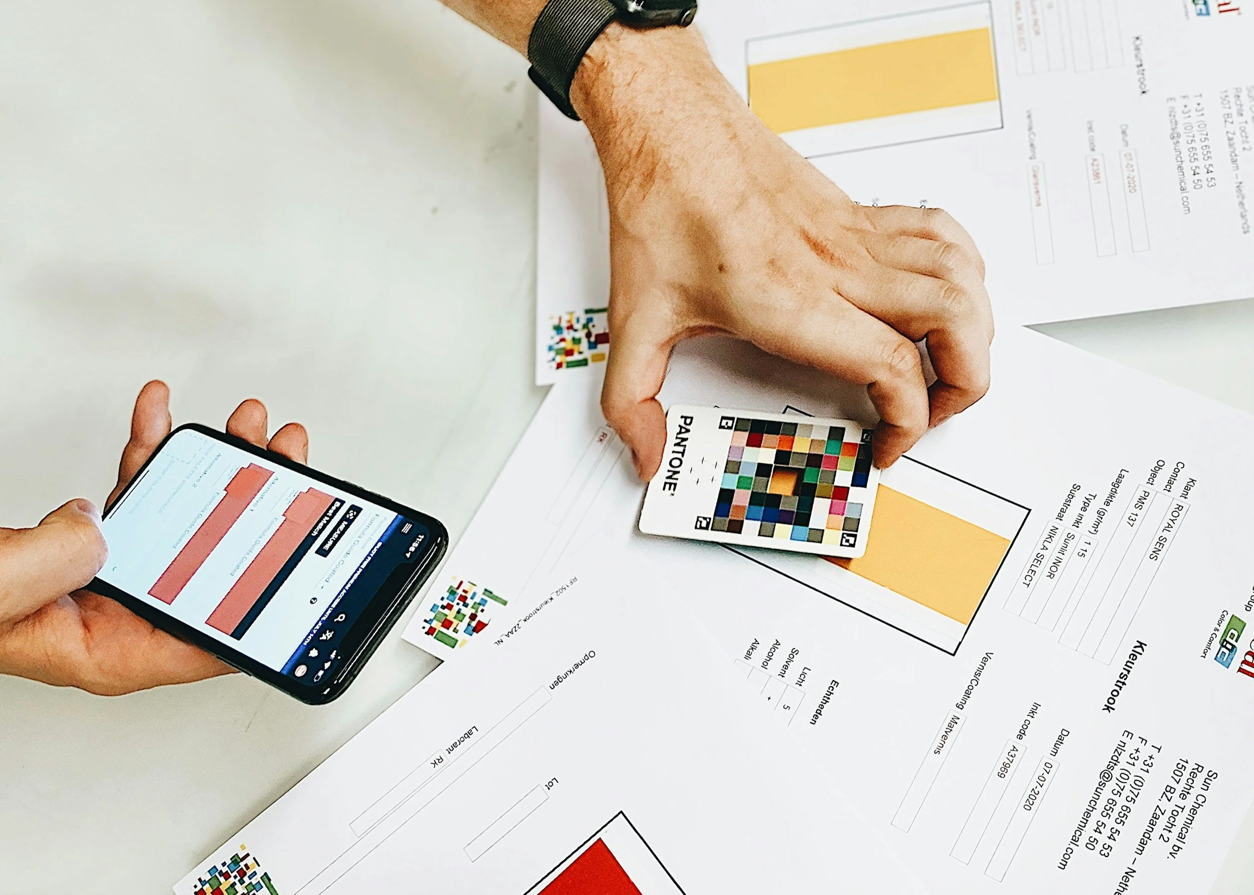A person using a smartphone and a color guide card during a color analysis or printing test, with printed color charts and information on a workspace.