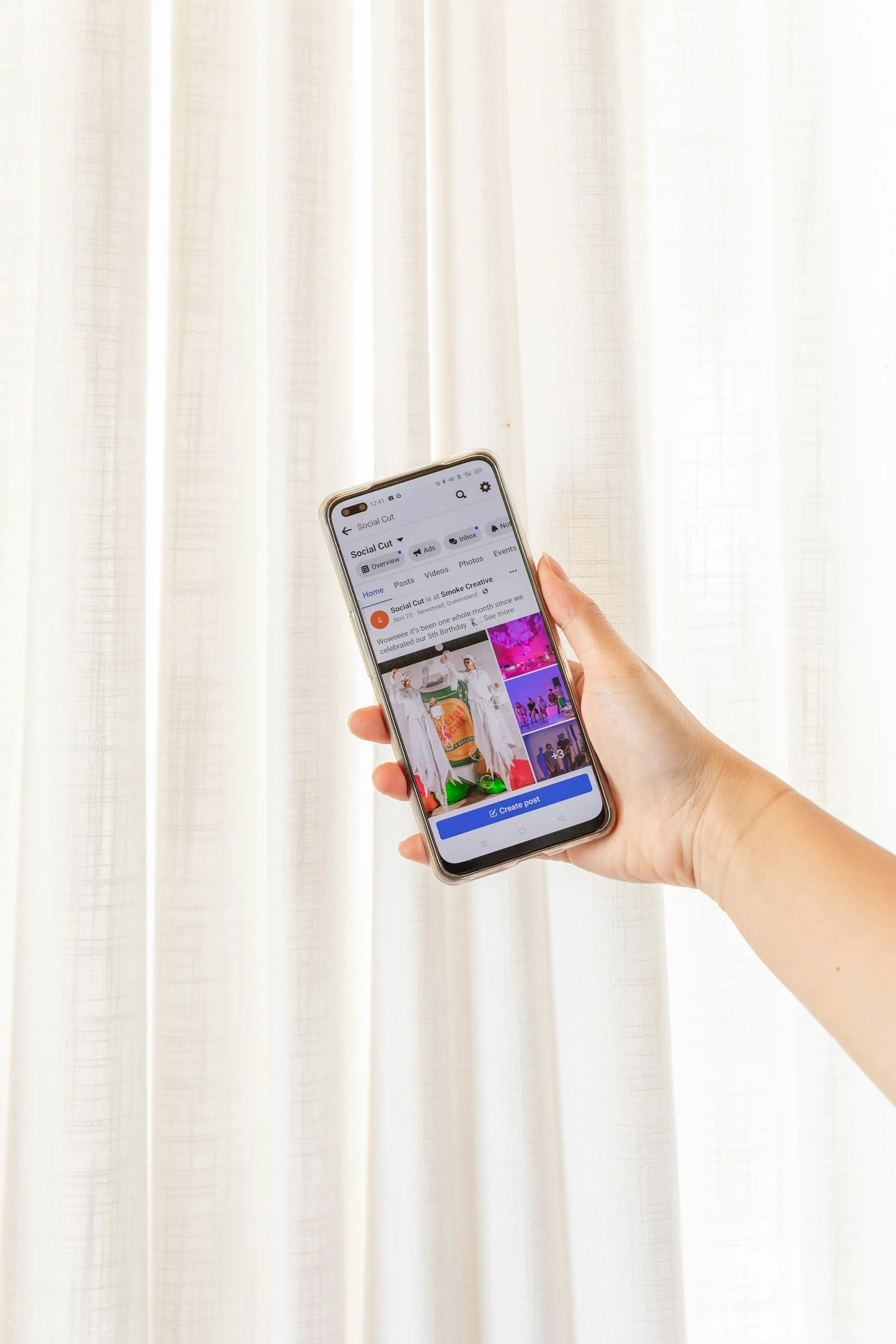 Hand holding a smartphone displaying a Facebook page titled 'Social Cut' in front of a light-colored curtain.