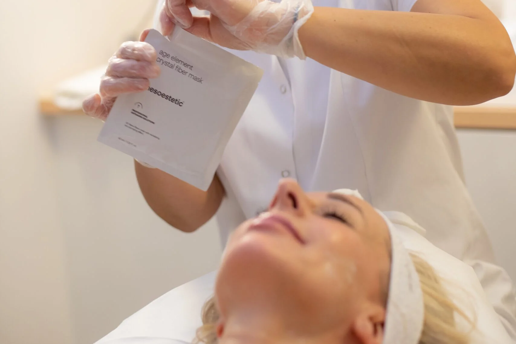 Una donna che riceve un trattamento estetico facciale, mentre un'estetista le mette in faccia una maschera a base di fibra di vetro, indossa guanti di plastica e un camice bianco.