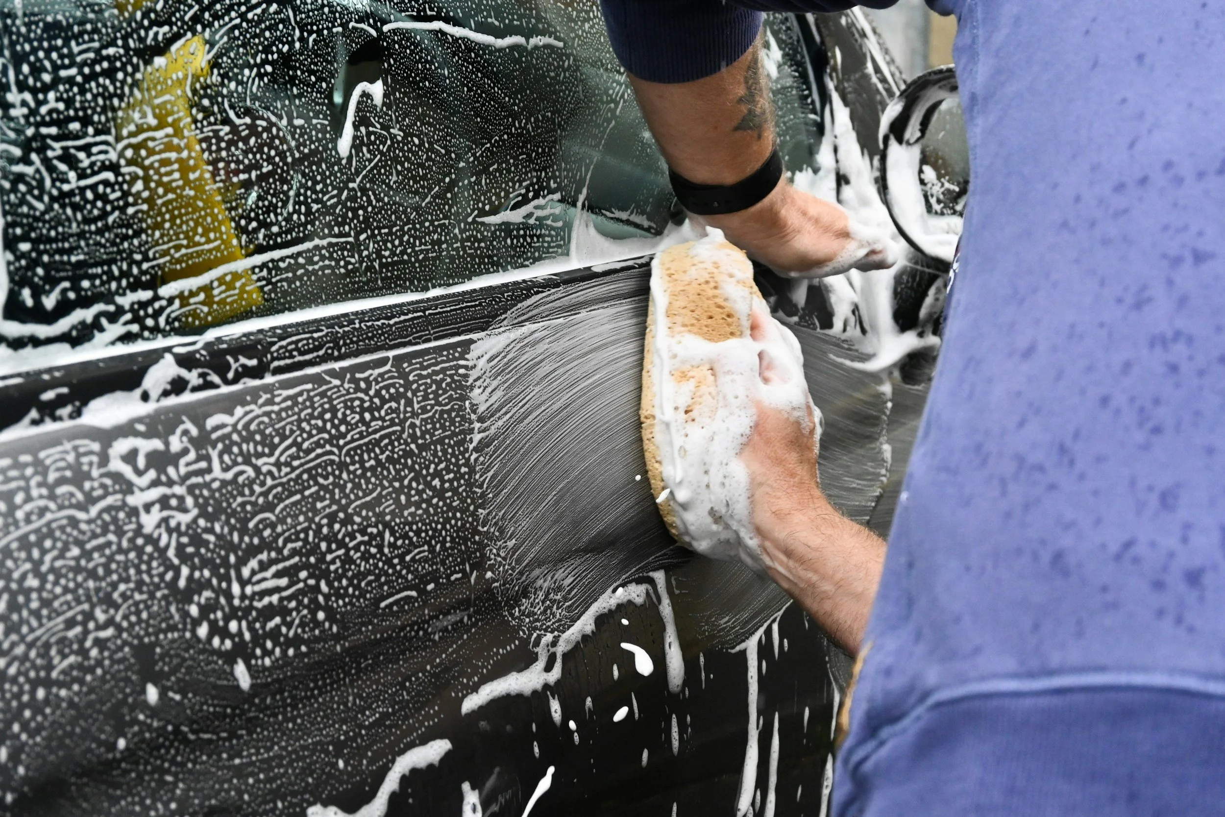Person washing a black car with a sponge, creating soap and foam on the surface.