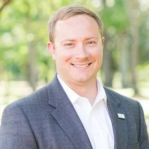 A smiling man with light hair wearing a gray blazer and white shirt standing outdoors in a park with green trees in the background.
