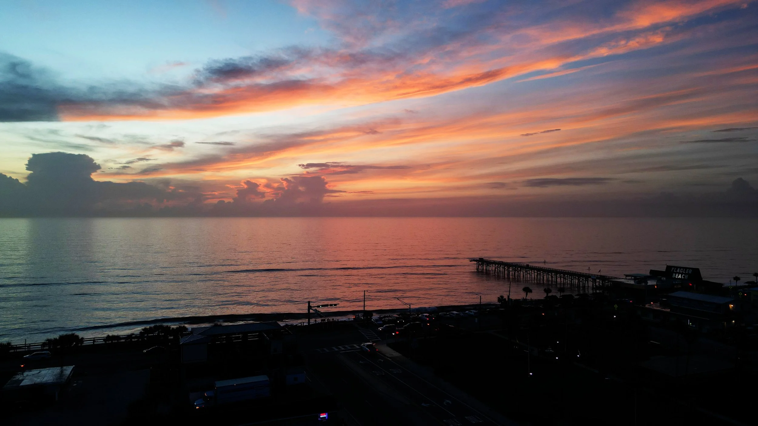 Sunset over the ocean with colorful sky and clouds, view from a building with a pier extending into the water, and the sign 'Flagler Beach' visible on a nearby building.