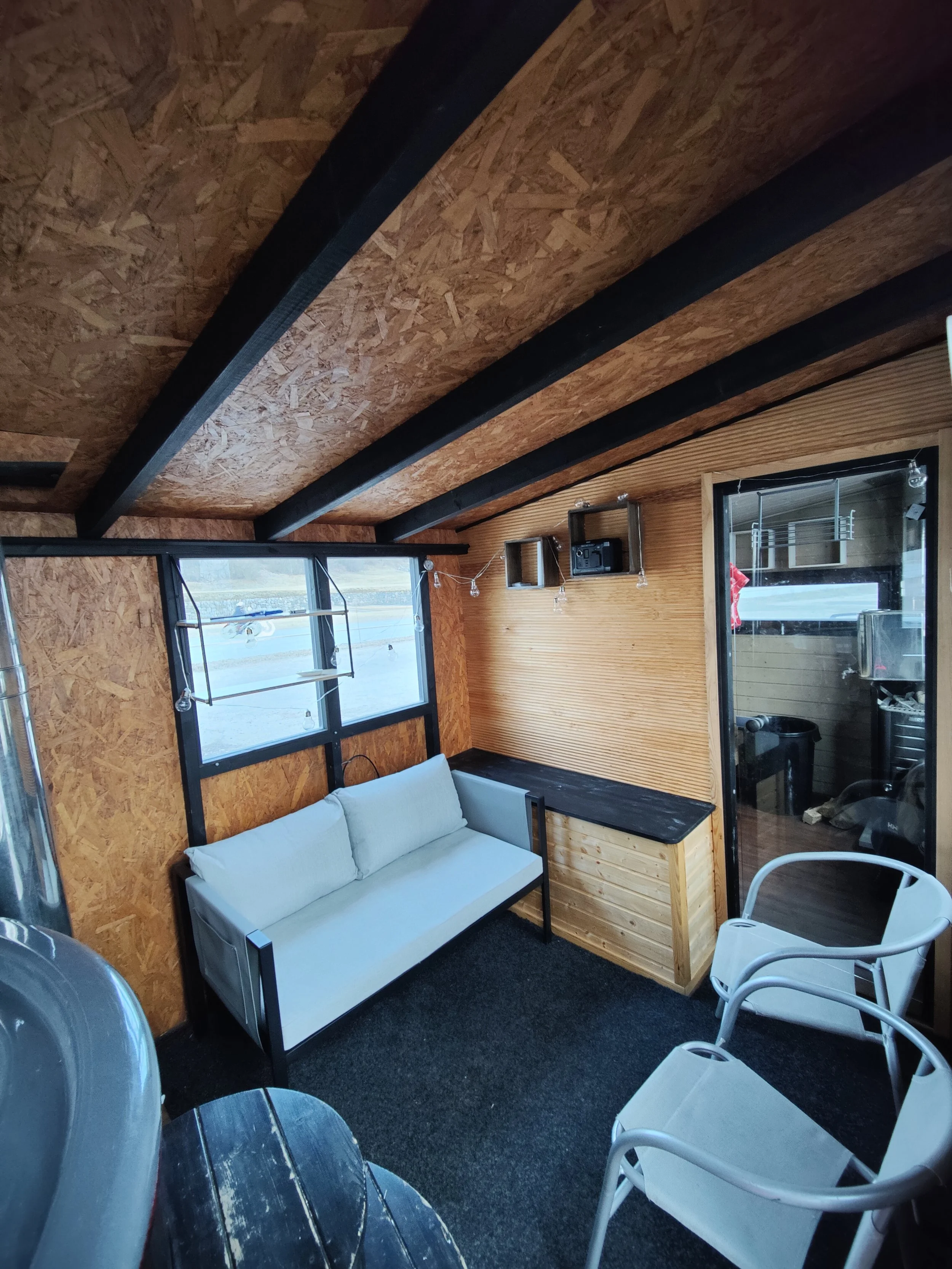 Sisäkuva puisesta mökkihuoneesta, jossa on valkoinen soffa, kaksi valkoista tuolia ja ikkuna. Seinät ja katto on paneelista ja katossa on mustat palkit. Tila on pieni ja viihtyisä.