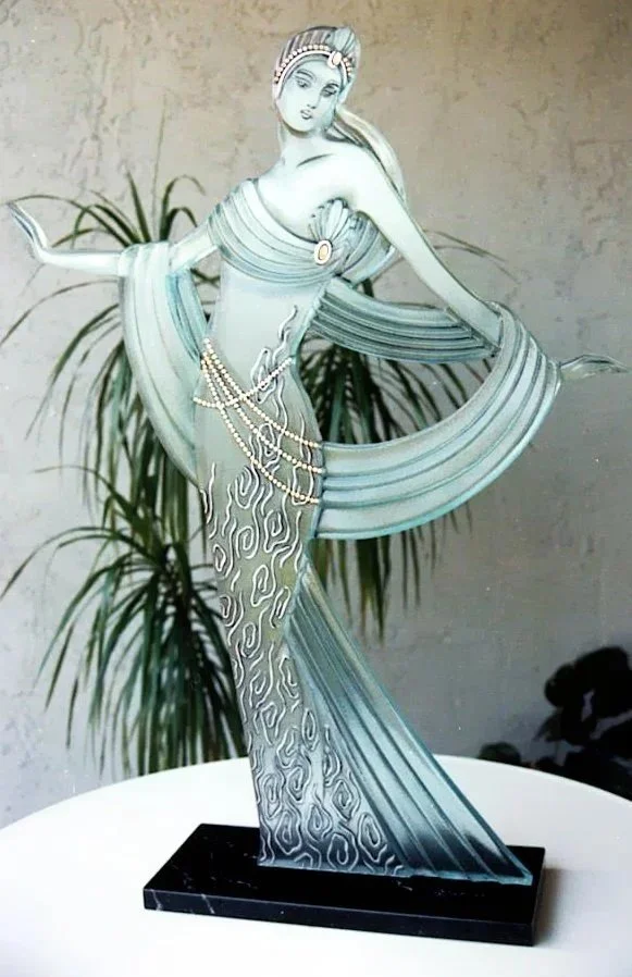 An art deco glass sculpture of a woman with flowing hair, dressed in a stylized gown and standing on a rectangular black marble base, with a background of potted plants and a textured wall.