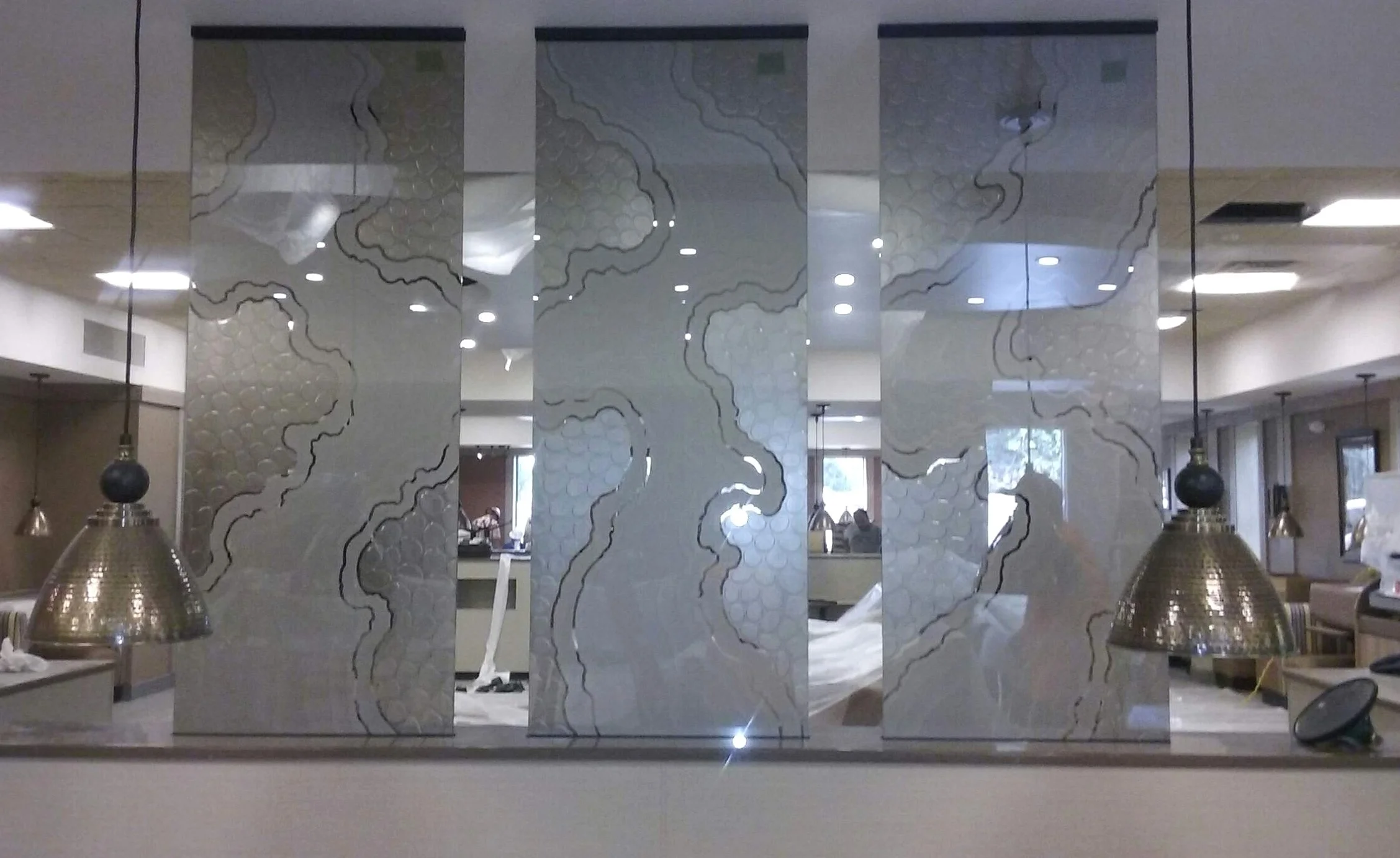 Interior of a restaurant with a decorative carved glass partition featuring an abstract pattern, pendant lights, and tables in the background.