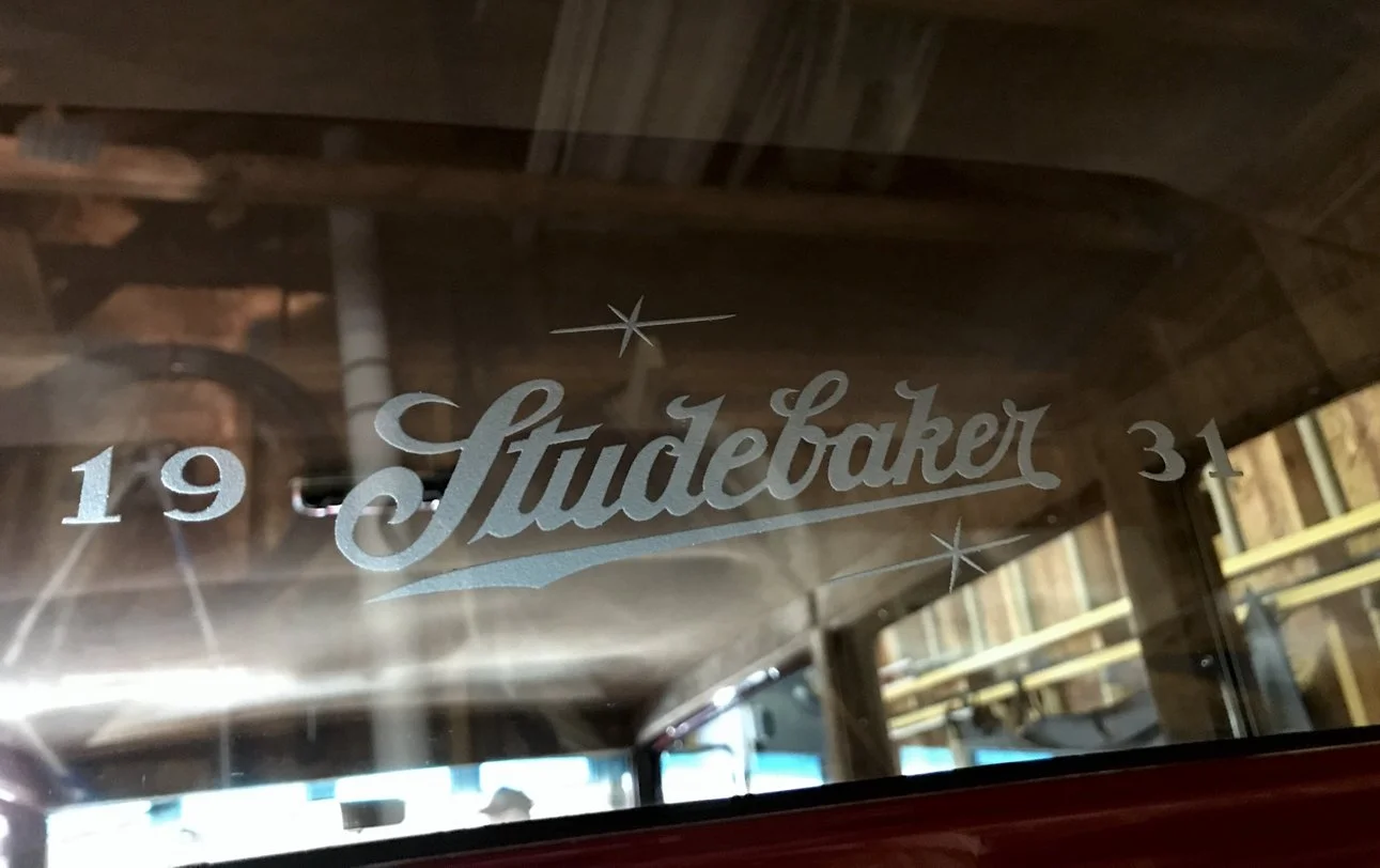 A glass window with glass etching that reads 'Studebaker' with the number '19' to the left and '31' to the right, and stars around the text, in a wooden garage or barn.